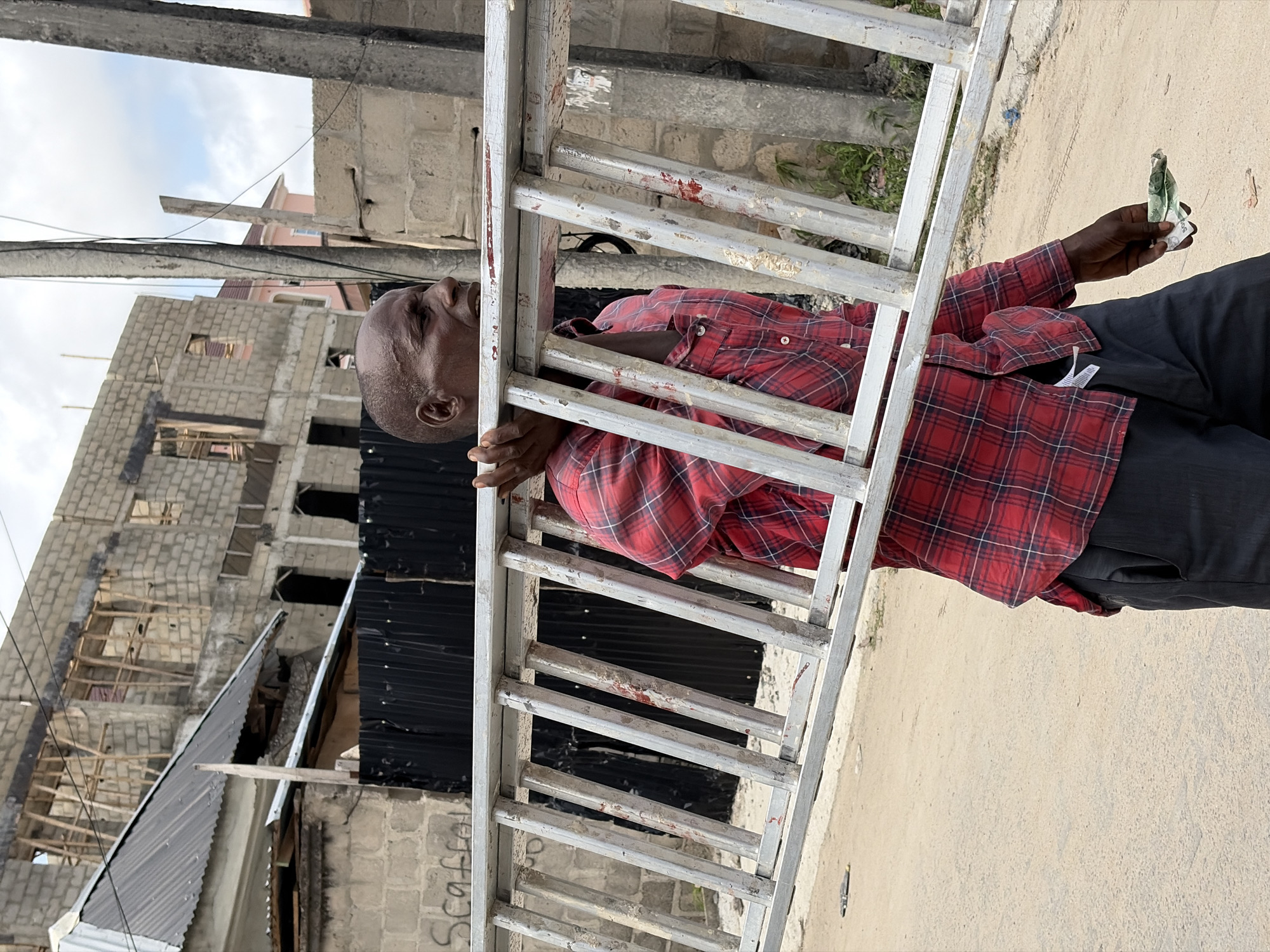 A man wearing a red plaid shirt and black pants is holding a ladder. The man is holding a wad of cash in his hand. The location is the city of Igboefon, Nigeria, as evidenced by the architecture. A building under construction is in the background.