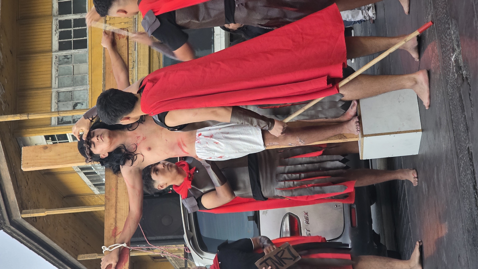 The image depicts a scene of the crucifixion of Jesus Christ, likely a Passion Play, in an unidentified urban environment. A man portraying Jesus is affixed to a wooden cross, with his hands bound by rope. He wears a crown of thorns, and there are simulated blood markings on his chest. Roman soldiers in costumes are present, one holding a sign that reads "INRI." A person is apparently fixing the crown of thorns. The scene is staged near a yellow building with multiple windows and in proximity to a silver car with "ST REX" visible on the side. A wooden box-like structure supports the base of the cross, and a wooden pole is also visible. The setting appears to be an outdoor public space.