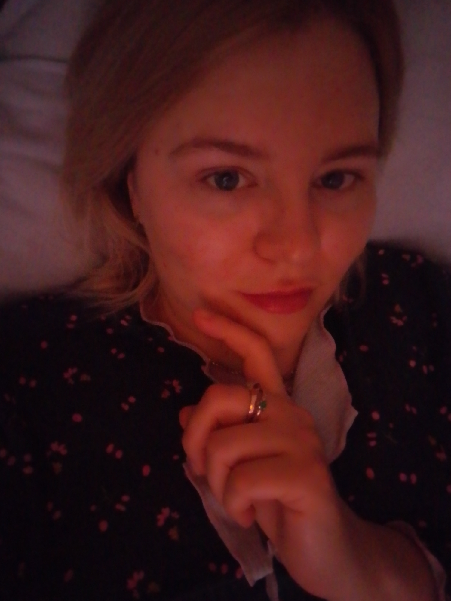 A fair-skinned woman with blond hair is lying down, possibly in bed. She wears a dark-colored top with a small, scattered floral pattern. Her left hand is raised to her chin, with a ring visible on one of her fingers. The lighting appears dim and reddish. There's no evidence to indicate that the location is in the city of Izhevsk, Russia.