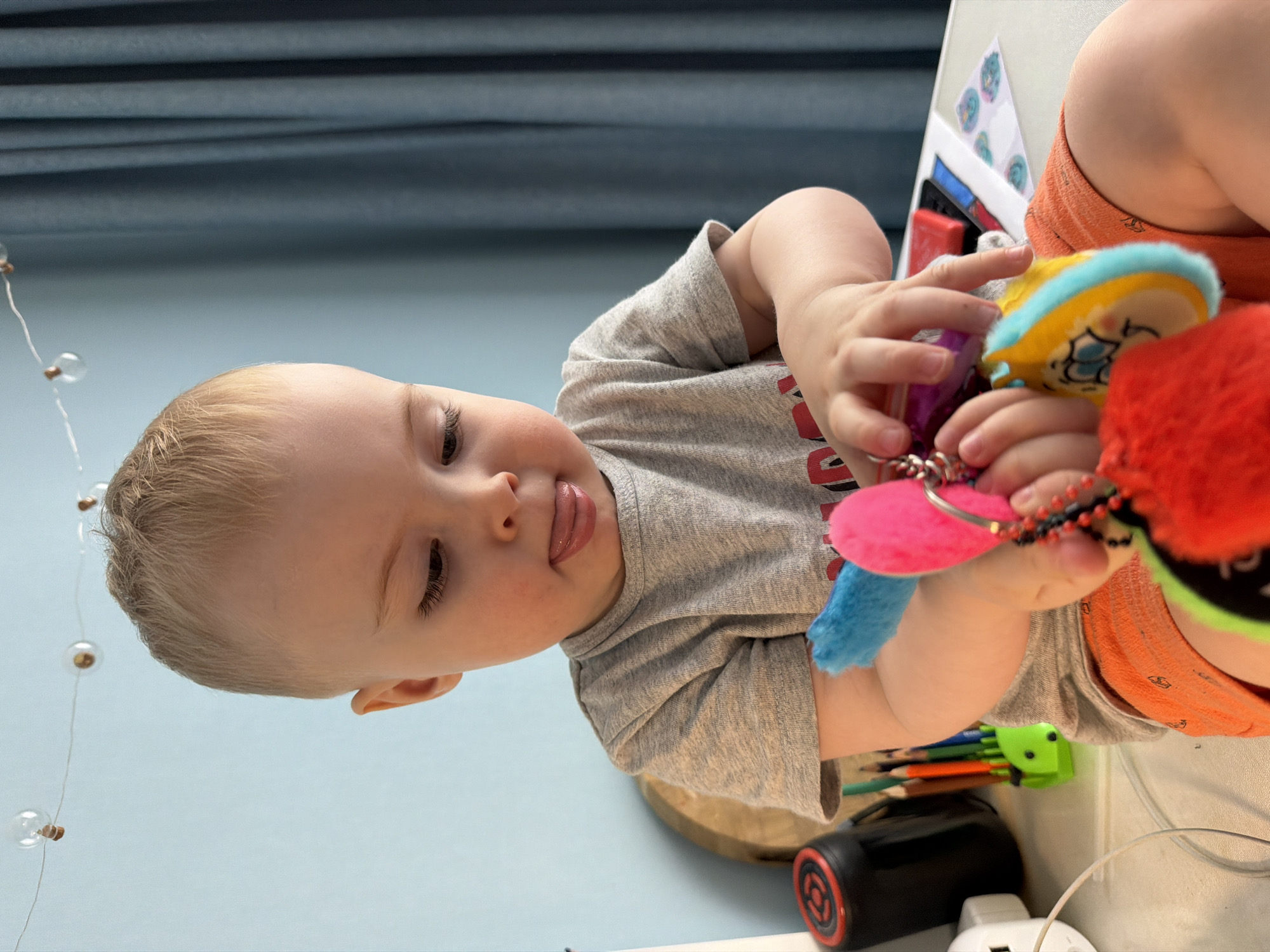 A close-up shot shows a fair-skinned toddler playing with colorful toys. The child, with light blonde hair, is wearing a gray t-shirt and orange pants. The toys appear to be a mix of fabric and plastic, including red, blue, pink, yellow, and green elements. The child's attention is focused on the toys held in their hands, and they are sticking their tongue out. The background is a mix of light blue and gray, possibly a wall and furniture. Near the child are art supplies, including a green pencil holder and various pencils. A round black object with red detailing and a white electrical outlet are also visible. The location is indeterminate, as the image provides no clues regarding Votkinsk, Russia or any other specific location.