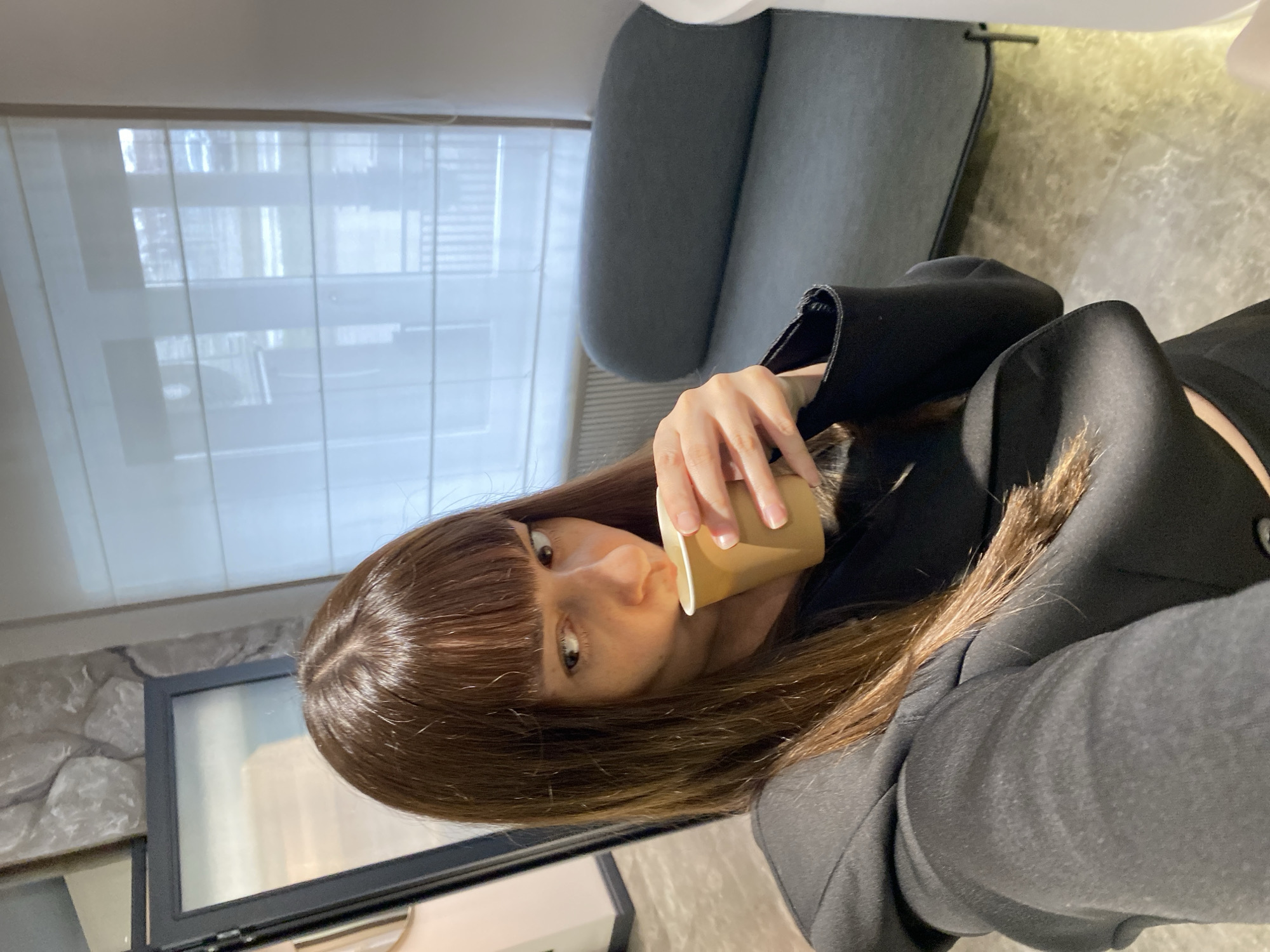 A person with brown hair and bangs is drinking from a beige cup. They are wearing a black blazer. A gray chair is visible behind them, and a window with white blinds is to the left. A stone-textured wall is partially visible. The location in Votkinsk, Russia, is unknown.