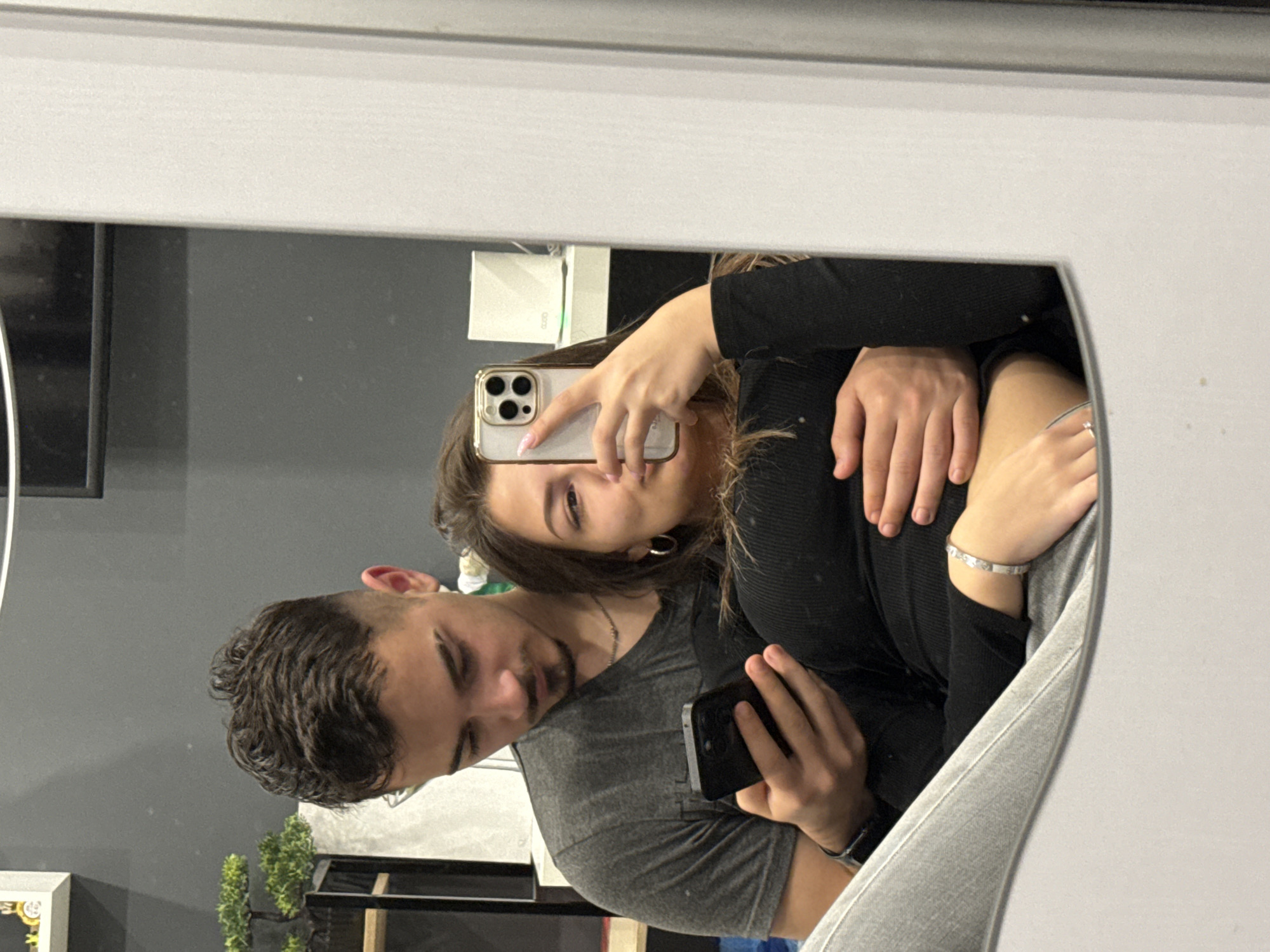 A young couple is taking a selfie in a mirror. The woman is holding a white iPhone with a triple-lens camera, while the man is holding a black smartphone. The woman is wearing a black long-sleeved top and has dark hair. She is also wearing earrings and a bracelet. The man has dark, curly hair and is wearing a gray t-shirt. A thin necklace is visible around his neck. He is also wearing a watch and a bracelet. The background shows a gray wall, a white shelf with a box, a television, and a potted plant. The couple is reflected in a rectangular mirror with a wide white frame. The location is potentially in an interior setting in the city of Sofia, Bulgaria.