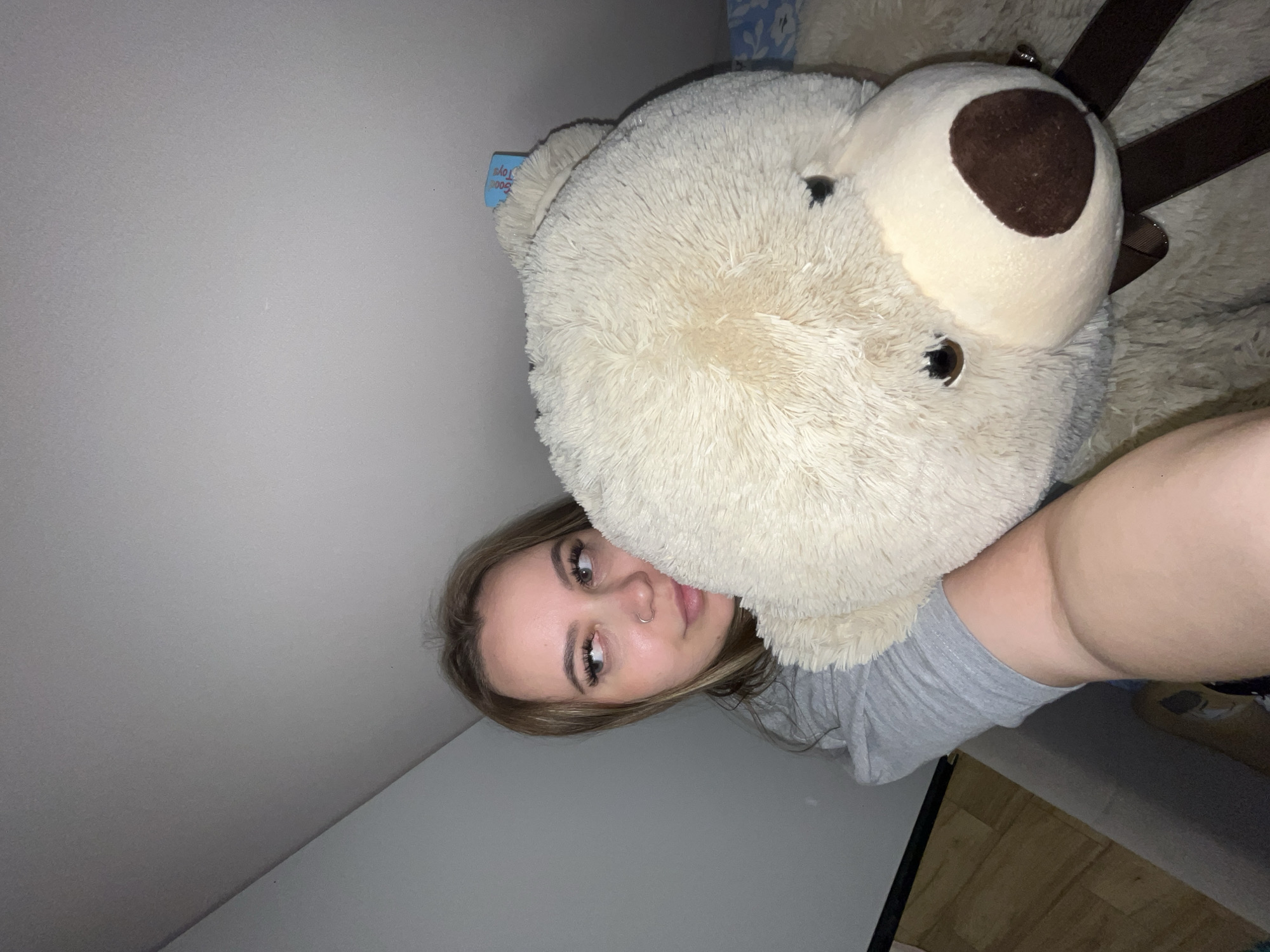 A young woman with light brown hair is posing for a selfie with a large, white teddy bear. The bear's nose is a darker brown color. The woman is wearing a gray shirt and has a nose ring. The background consists of a gray wall and ceiling. The location is not discernible from the visual information provided.