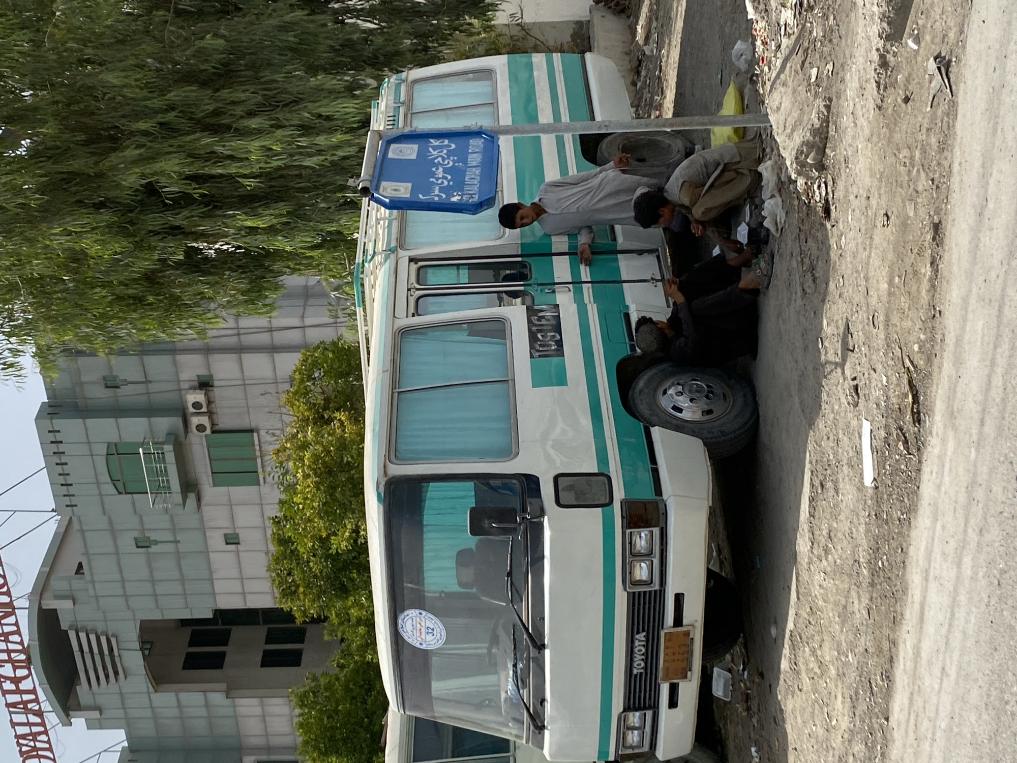 The image shows a white and teal colored Toyota Coaster bus parked on a dirt road in Kandahar, Afghanistan. The bus has the word "TOYOTA" written on the front grille. A blue signpost is next to the bus, which has writing in both Arabic and English that reads "KALADCHA MAIN ROAD". The building behind the bus appears to be a modern multi-story structure with tinted windows and the partially visible text "ROYAL AFGHANISTAN" on its facade.
In the foreground, three young boys are sitting on the ground near the bus. One boy, wearing a light grey shalwar kameez, is standing next to the bus door, holding what appears to be a cigarette. Two other boys are seated on the ground by the bus's rear wheel. One is wearing dark clothing, and the other is wearing a light brown shalwar kameez. They are surrounded by scattered debris, including plastic bags and discarded paper. The ground is a mix of dirt and gravel. A large tree with drooping branches stands behind the bus.