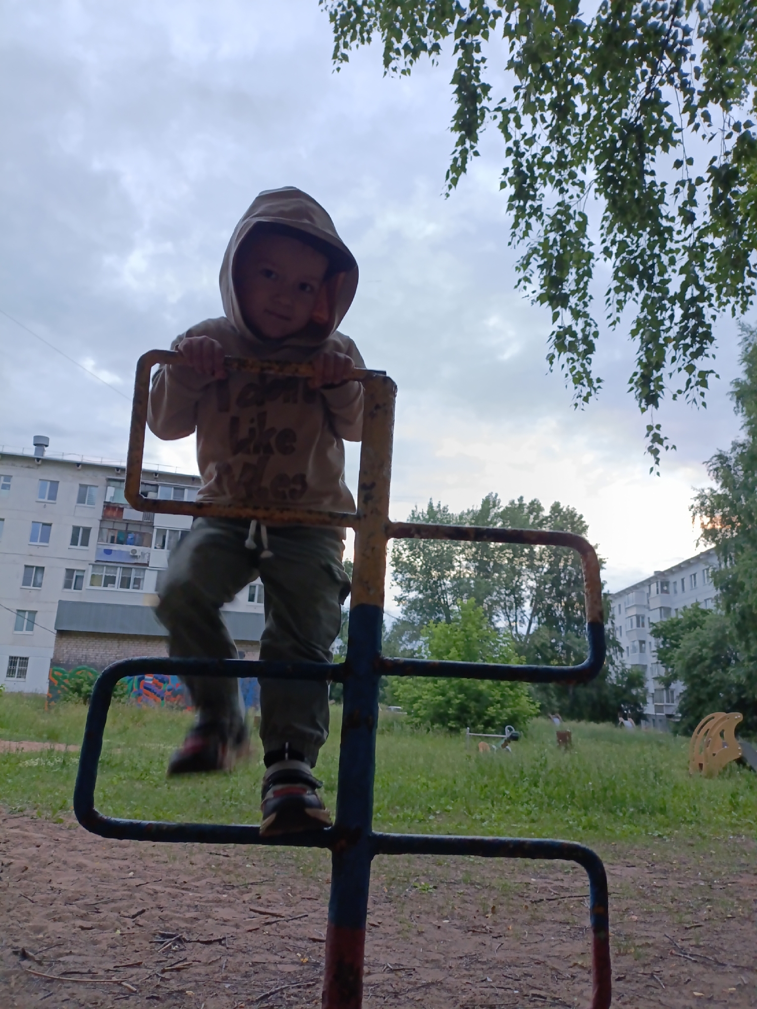 A low-angle outdoor shot captures a young child, wearing a hooded sweatshirt and green pants, climbing on a rusty blue and yellow metal playground structure. The child is looking towards the camera. In the background, a large, multi-story apartment building with white siding and visible balconies is situated to the left. A grassy area with trees and other playground equipment, including a yellow slide, can be seen behind the metal structure. The sky is overcast with clouds. The scene appears to be in a residential area of Chaikovsky, Russia, likely a courtyard playground.