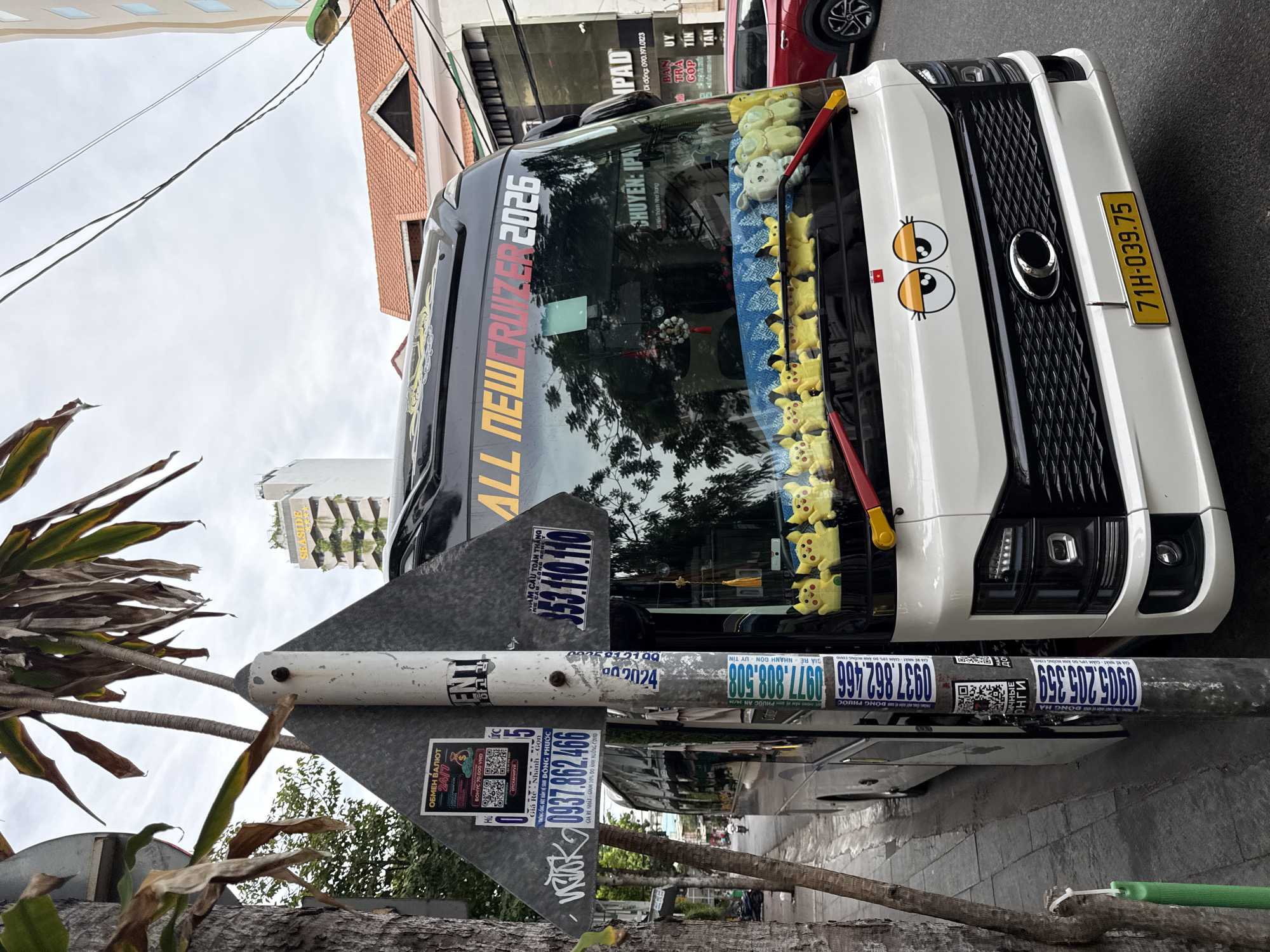 The image shows the front of a white bus with a black grille and yellow license plate. The bus is parked on the right side of a street in Nha Trang, Vietnam, with a sidewalk and trees on the left. The bus has "ALL NEWCRUZER 2026" written on the windshield. There are several yellow stuffed Pikachu toys and some white stuffed animals lined up on the dashboard. A cartoon face with large eyes is stickered on the back of the bus. A metal pole with various stickers is in the foreground, to the left of the bus. In the background, there is a multi-story building with red tile roofs and a taller building with a sign that reads "SEASIDE HOTEL". The sky is overcast.