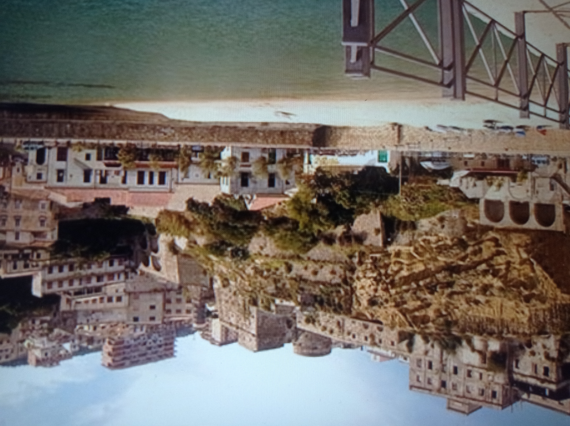 This is a medium shot of a coastal town with buildings perched on a steep, rocky cliff face. The buildings are a mix of old stone structures and more modern constructions, many with terracotta roofs. Some buildings appear to be partially abandoned or in disrepair. In the foreground, there is a sandy beach with a stone wall and a metal railing. Several small boats are pulled up onto the sand. The sea is a calm, green color. The sky is a clear blue with a few wispy clouds. The scene depicts a sunny day in a picturesque but weathered seaside location.