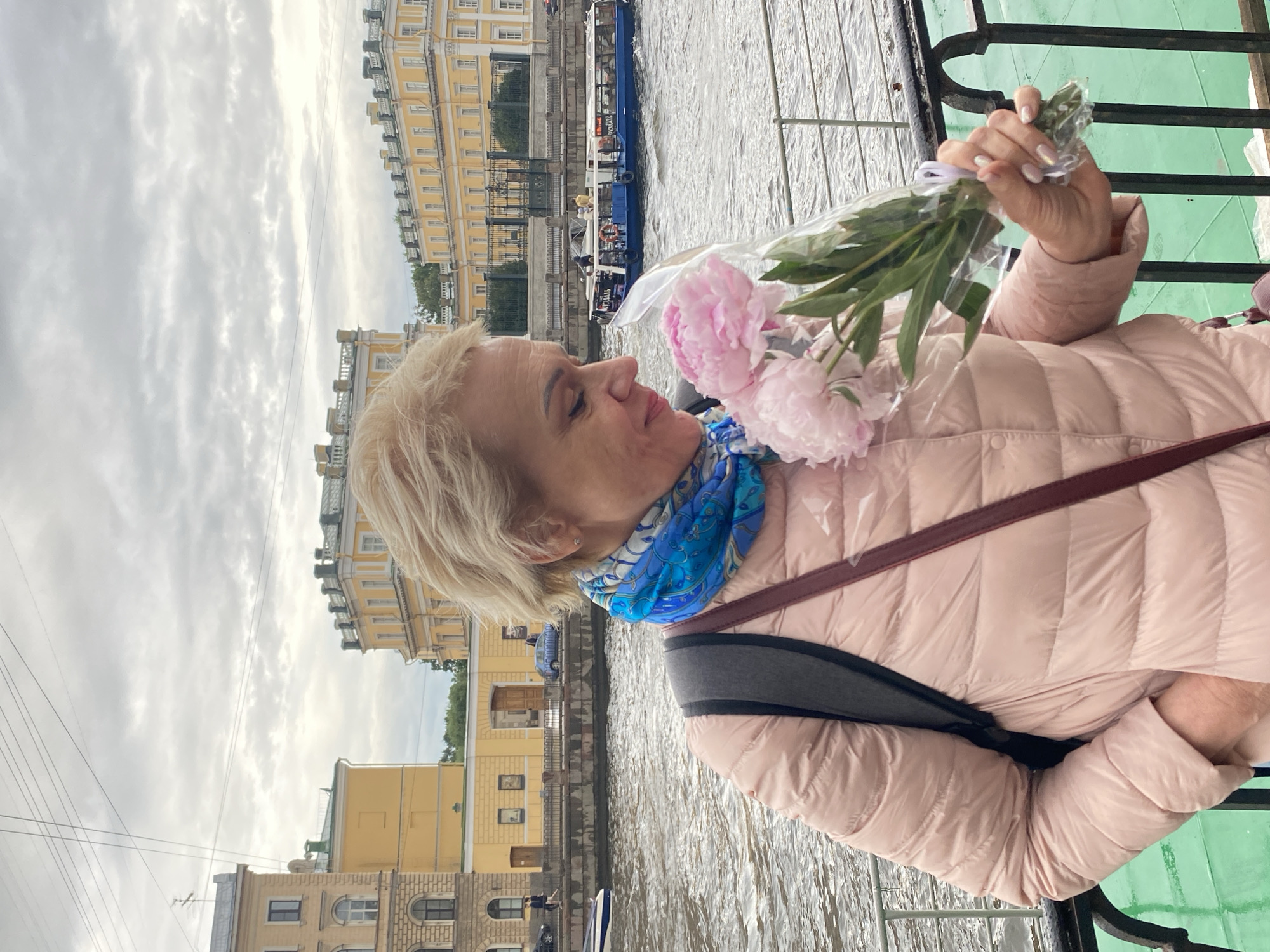 A woman with blonde hair is holding a bouquet of pink peonies, wrapped in cellophane, while standing on a pier in Saint Petersburg, Russia. She is wearing a light pink puffer jacket and a blue and white scarf. Behind her, a canal or river flows, with boats on the water and historic yellow buildings lining the embankment. The sky is overcast with clouds. The woman appears to be looking down at the flowers.