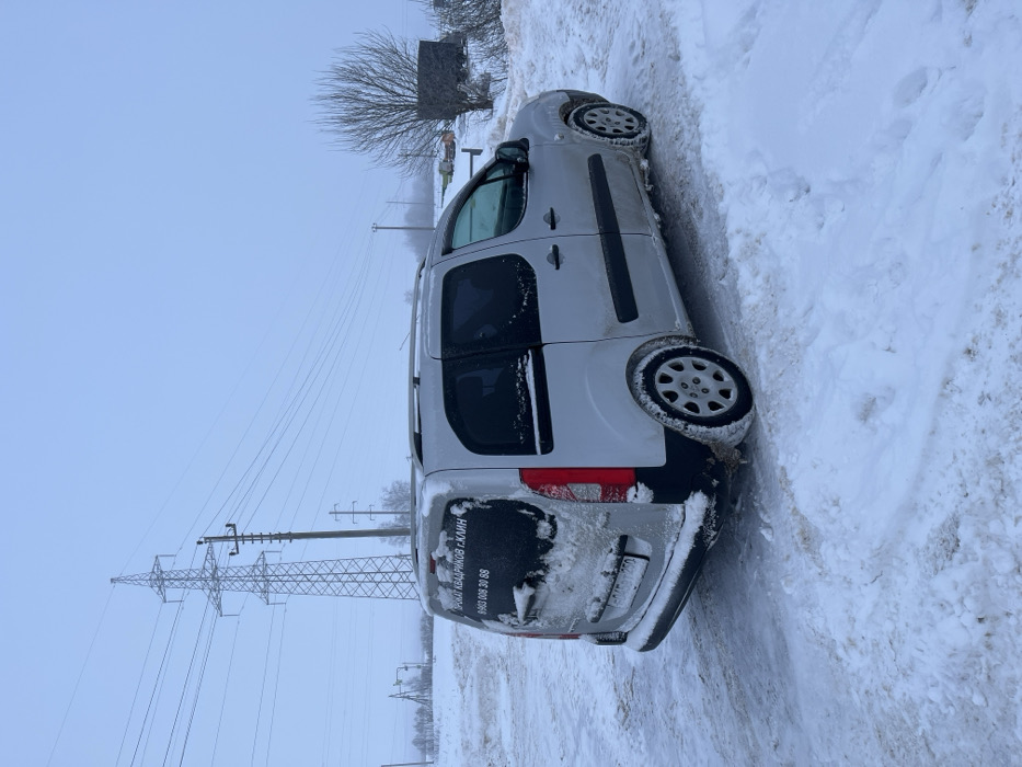A light-colored van, possibly silver, is parked on a snow-covered roadside in Voronino, Russia, under an overcast sky. The vehicle is angled with its rear left side facing the viewer, partially covered in snow and slush. Cyrillic text on the rear window reads "ПРОКАТ КВАДРИКОВ Г. КЛИН" (Quad Bike Rental Klin City) followed by a phone number "8 903 008 30 88". A Russian license plate, "A 494 YX 750", is affixed to the rear. In the background, a large metal lattice electrical transmission tower and several utility poles support multiple power lines extending across the winter landscape. The ground is uniformly covered in snow, with visible tire tracks on the unpaved surface. To the right, a leafless tree and a dark, low structure are partially obscured by snow. No individuals are present in the scene.