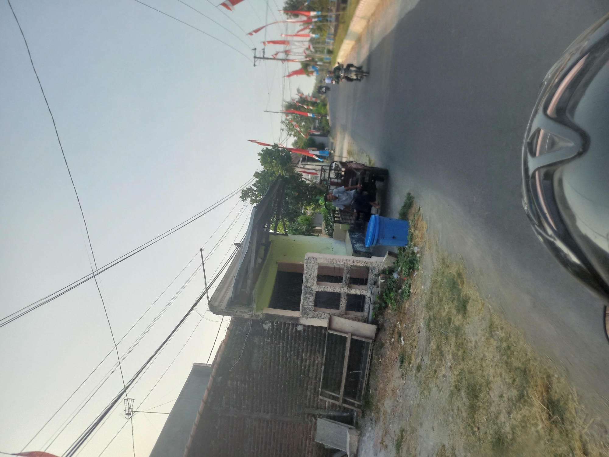 The image depicts a street scene in Kedung Otok, Indonesia. A man is seated on a red cart pulled by a vehicle. In the background, several motorcycles are visible on the road, some with riders. The street is lined with numerous red and white flags, indicating a festive or commemorative occasion. To the left of the road, a brick building with a green-colored facade is present, along with a blue trash bin. The sky is clear and bright. The perspective suggests the photo was taken from the viewpoint of someone riding a motorcycle, as the front of a helmet is visible in the foreground.
