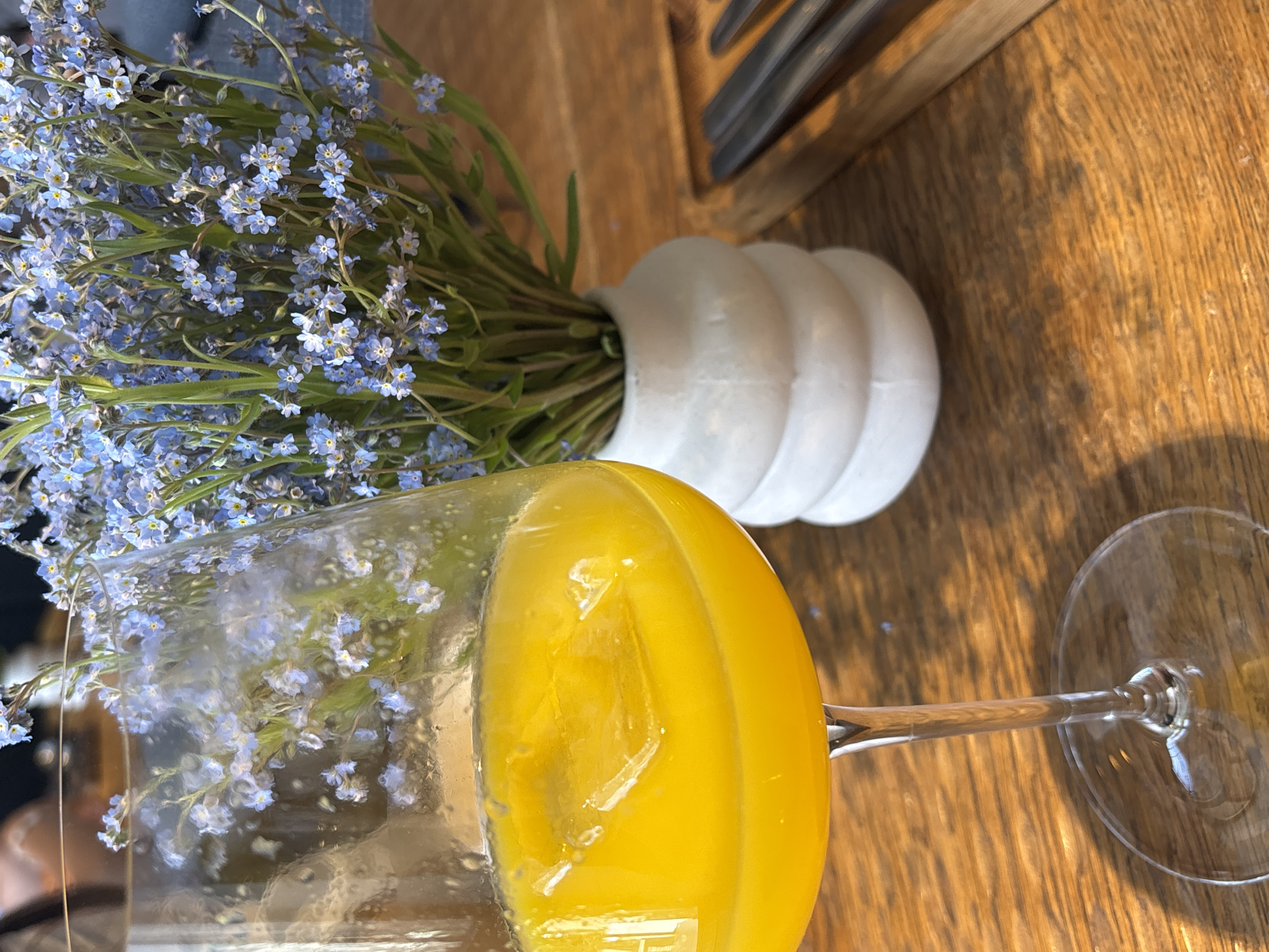A bright, close-up indoor photograph captures a table setting in Krasnoyarsk, Russia. In the immediate foreground on the left, a clear, stemmed glass holds a vibrant yellow liquid, likely juice or a cocktail, with several partially submerged ice cubes. To the right of the glass and slightly further back, a small, white, ribbed ceramic vase contains a dense bouquet of delicate blue forget-me-not flowers with small yellow centers and green stems. All objects rest on a polished, light brown wooden table with a visible horizontal grain. In the upper right corner, a portion of metallic cutlery, appearing to be a knife and fork, is visible on a wooden board. The background is softly blurred, showing indistinct shapes and muted tones consistent with an occupied indoor dining environment. No individuals are clearly discernible.