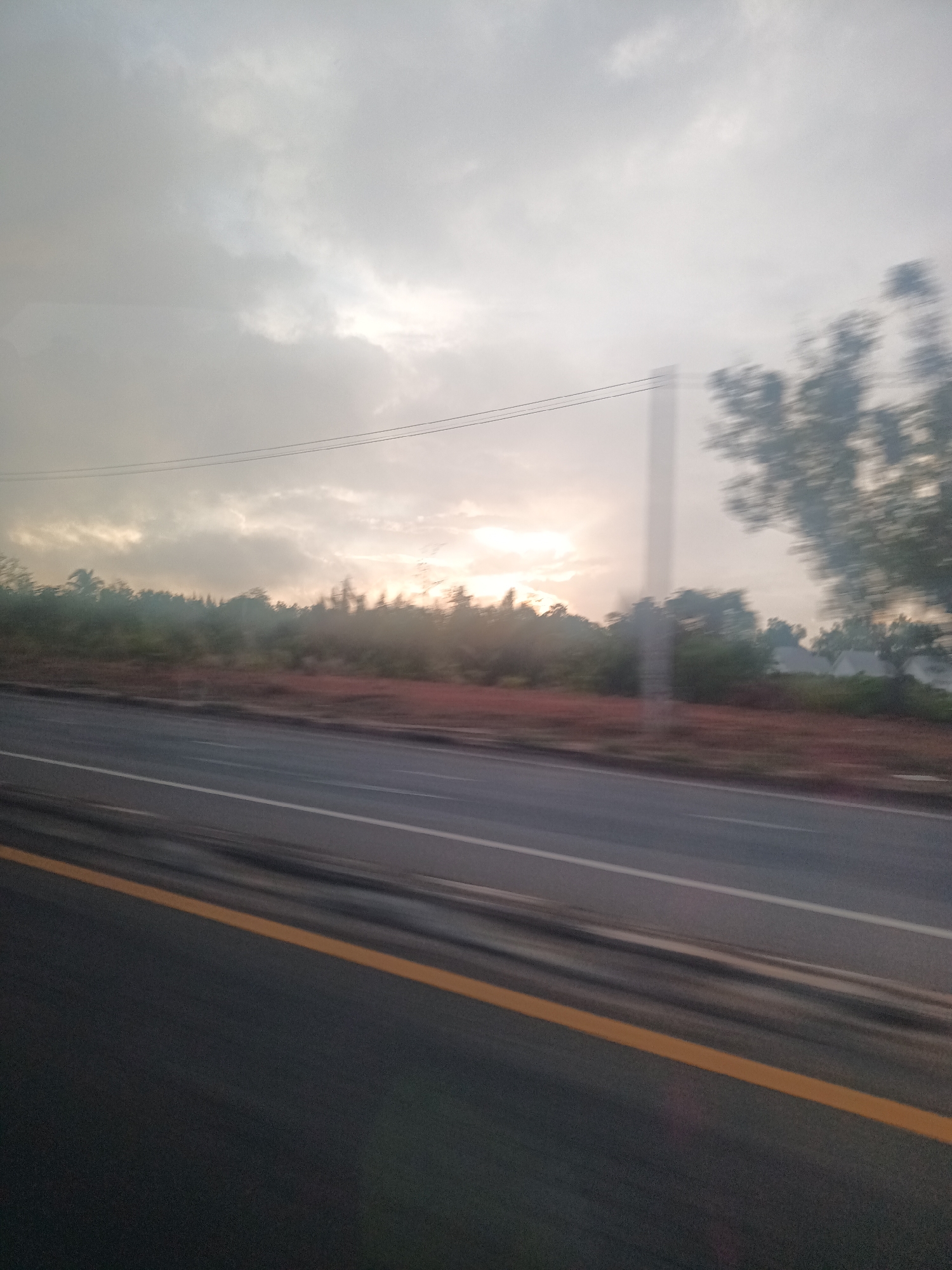 The photograph appears to be taken from a moving vehicle on a road in Surat Thani City Municipality, Thailand. The road is paved with a yellow line on the side. There's vegetation on the roadside with trees and foliage. The sky is overcast, with some sunlight breaking through the clouds. There are power lines running above the trees. The image has motion blur.