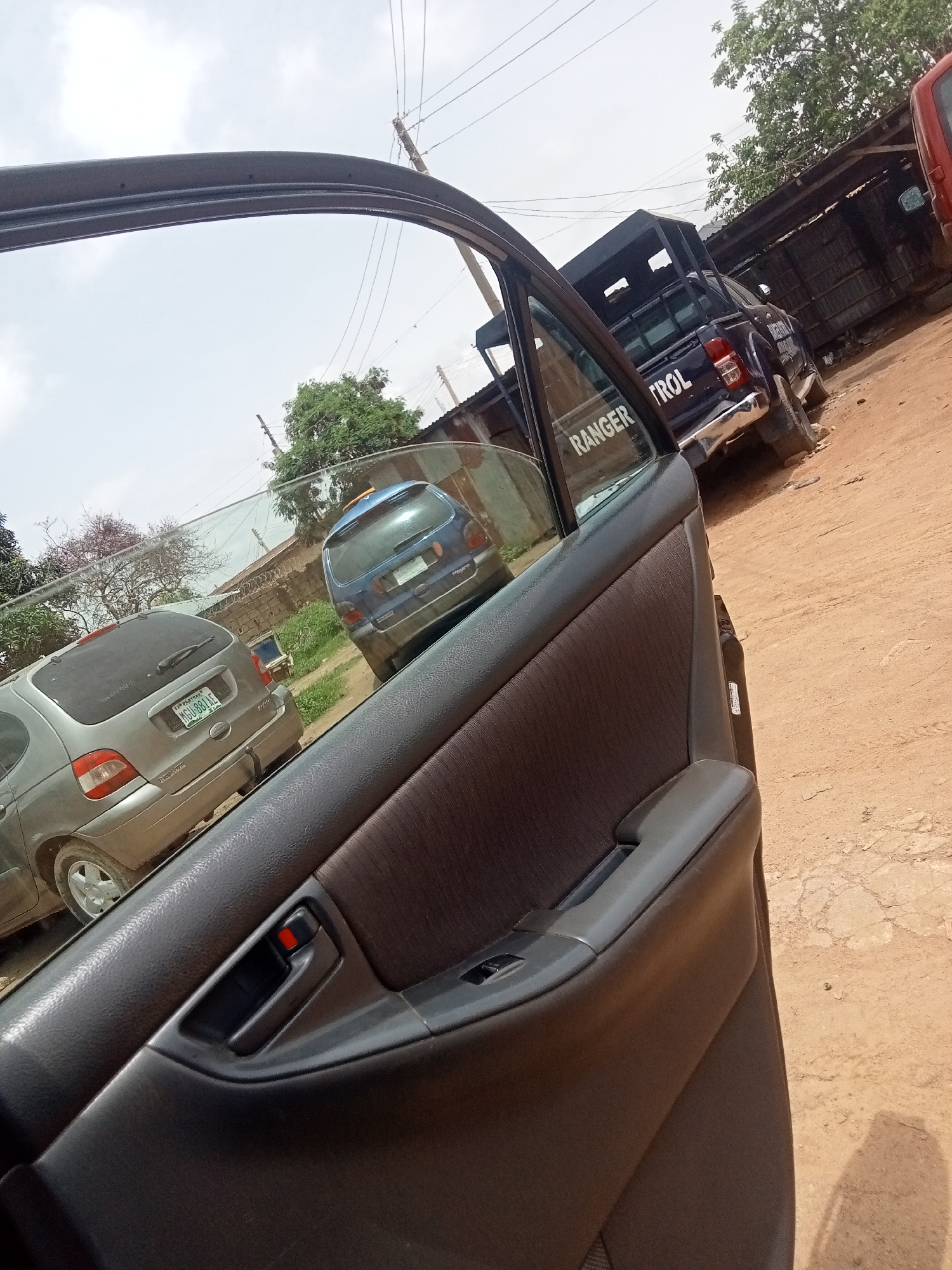The image shows a car door, with other cars parked nearby. A gray Renault Scenic is visible through the car window. The environment is characterized by dirt roads, suggesting the image was taken in Rafin Par, Nigeria. A "Ranger Patrol" vehicle is also present. There are no visible people.