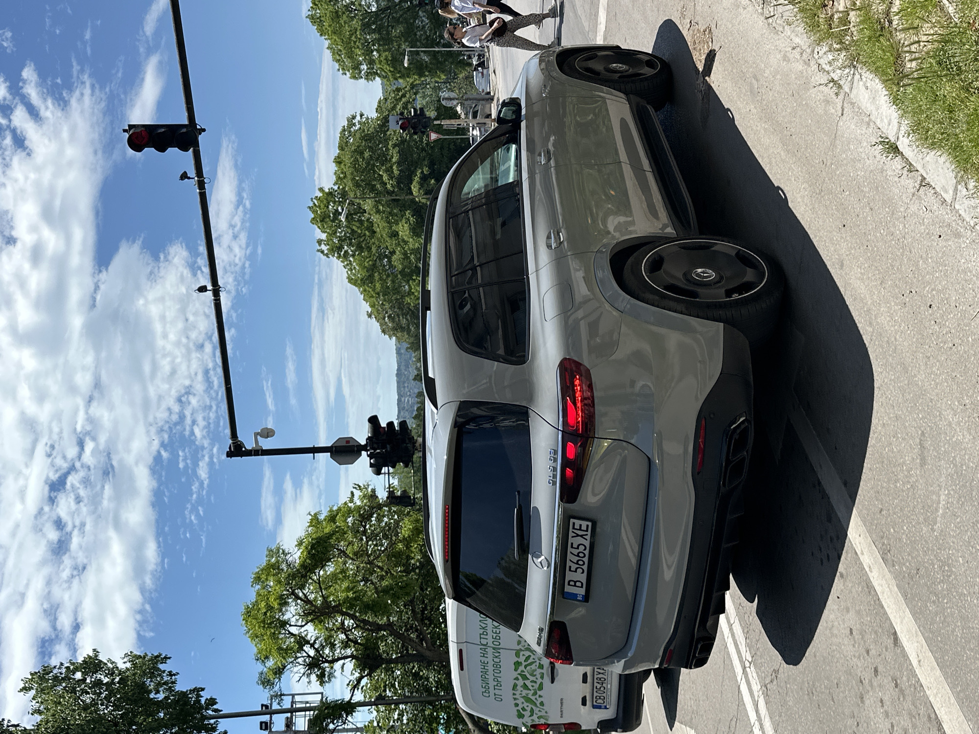 The image depicts a gray Mercedes-Benz GLC SUV stopped at a traffic light in Varna, Bulgaria. The car's license plate is "B 5665 XE". A white van, partially visible behind the SUV, is marked with green leafy graphics and bears the license plate "CB 0548 XA". The traffic light is showing a red light. Trees and buildings are visible in the background. Two pedestrians are crossing the road further up the street. The sky is blue with scattered white clouds. The scene appears to be taken during the daytime.