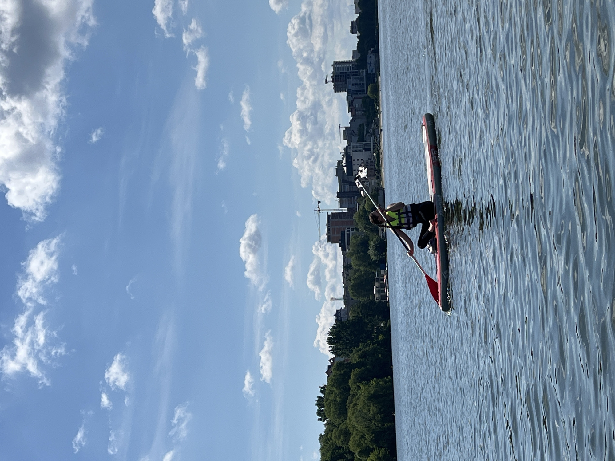A person is paddleboarding on a body of water, with a city skyline visible in the background. The person is wearing a life vest and is holding a paddle. The water is calm, and the sky is blue with some white clouds. The city skyline consists of buildings and trees. This scene appears to be in Bila Tserkva, Ukraine, a city known for its natural beauty and proximity to rivers and lakes.