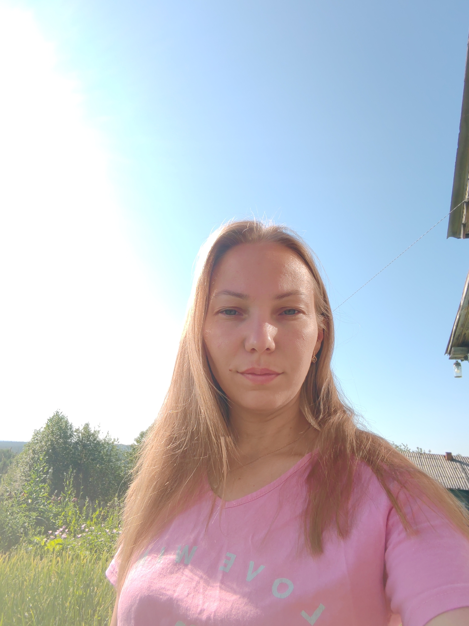 A selfie taken outdoors in daylight, likely in a rural area, possibly in Чухлэм, Russia. The subject is a fair-skinned woman with long, blonde hair, wearing a pink t-shirt with light blue lettering that appears to spell out "LOVE". She has light blue eyes and is looking directly at the camera. The background includes a bright sky, green trees, and tall grass. To the right, a portion of a wooden structure with a roof and a hanging lamp is visible. The sun is high in the sky, casting a bright light.