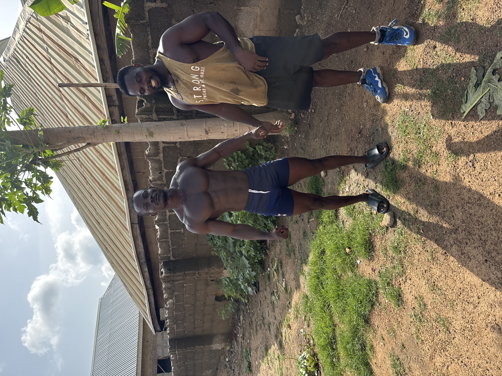 Two muscular men are standing outdoors, seemingly posing for a photo. One man is wearing a tan tank top with text, dark shorts, and blue athletic shoes. He is smiling and appears to be slightly bent at the waist. The other man is shirtless, wearing dark blue shorts, and sandals. They are holding hands. The background includes a corrugated metal roof, a brick wall, a tree, grass, and dirt. The location seems to be Akwanga, Nigeria.