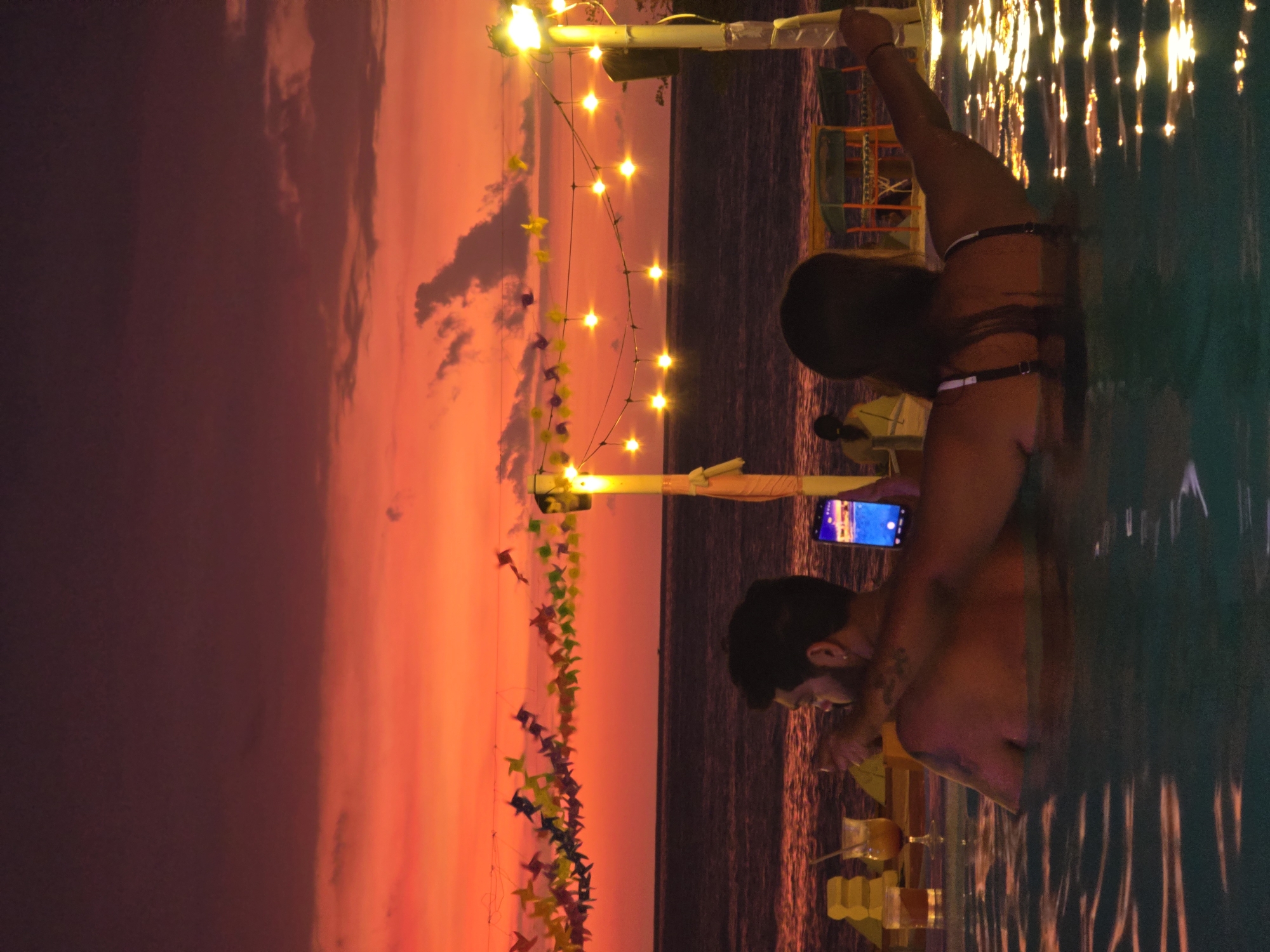 The image shows two people in a pool at dusk. The sky is a vibrant red and orange. There are string lights and colorful flags hanging above the pool. The people appear to be a couple; the man is holding a drink and the woman is looking at her phone. The location is unknown.