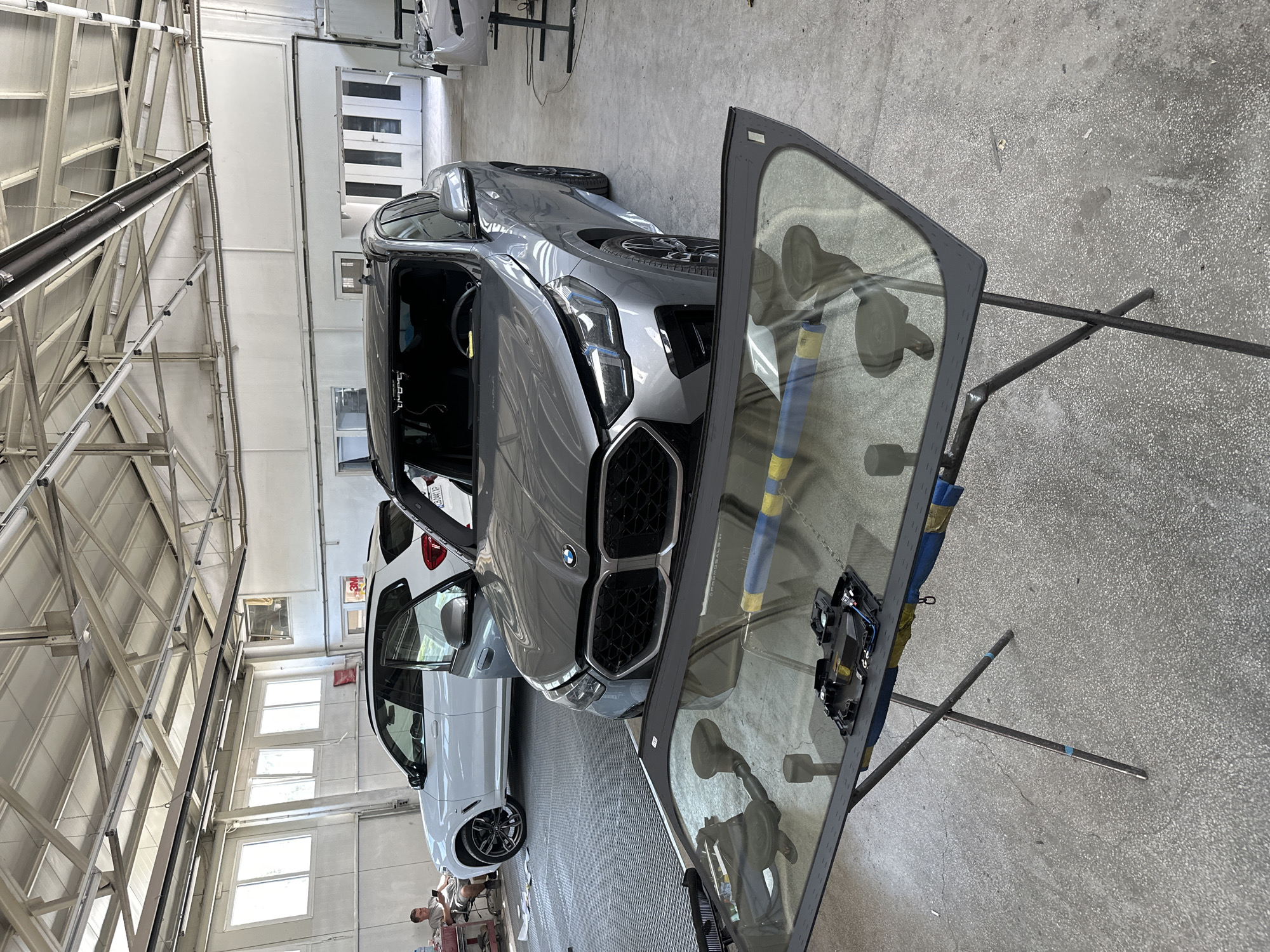 An indoor, well-lit garage scene features two SUVs and a windshield laid out on a portable stand in the foreground. The garage has a high, industrial-style ceiling with exposed beams and fluorescent lighting. The floor is concrete.
In the center, a gray BMW SUV is parked, its windshield removed. To its left, a white BMW SUV is also parked, with its driver's side door open.
In the foreground, a detached windshield, likely from the gray BMW, rests on a metal, X-shaped stand. The windshield has a black trim and suction cups attached. A piece of electronic equipment and blue and yellow tape are visible on the windshield.
To the far left of the image, a person is seated on a red toolbox, looking at a mobile phone. The background shows large, white doors, possibly leading outside, and another vehicle in a bay.