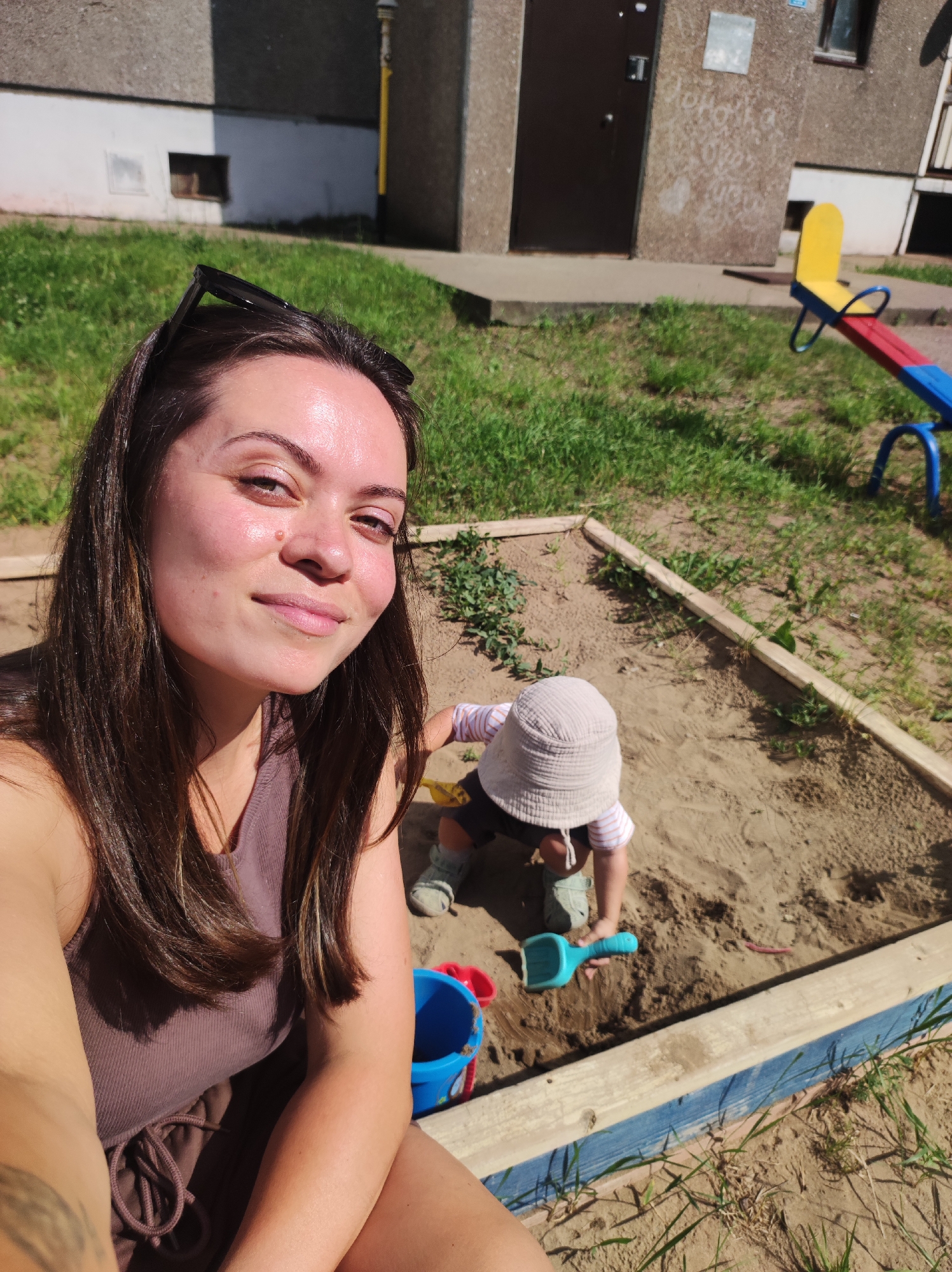 A woman and a toddler are in a sandbox in an outdoor play area. The woman is in the foreground, taking a selfie. She has long, dark brown hair and is wearing a purple tank top and matching shorts. Her sunglasses are on top of her head. The toddler, wearing a light-colored sun hat, is in the background, playing in the sand with a blue shovel. A red and blue seesaw is visible in the background, along with a concrete building and grassy area. The sandbox has wooden borders. The scene appears to be in a residential area.