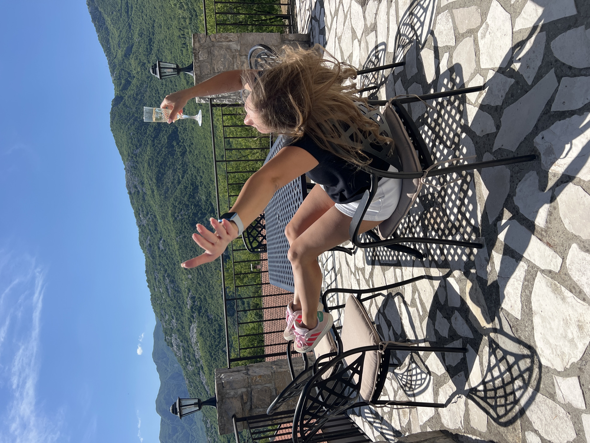 The image depicts a young woman seated on an outdoor patio, enjoying a scenic view under bright daylight. She has long, light brown hair and is wearing a black t-shirt, white shorts, and pink athletic shoes. Her right hand extends outwards, holding a champagne flute filled with a sparkling beverage, while her left hand is also outstretched.
She is seated at a dark metal outdoor table with a lattice design, on a matching chair with a light cushion. The patio floor is made of irregular, light-colored stone tiles. Strong sunlight casts prominent, intricate shadows from the furniture onto the ground, indicating it is likely mid-day or afternoon on a very sunny day.
The background reveals a magnificent mountainous landscape, densely covered with green trees, stretching under a clear blue sky with a few subtle clouds. A dark metal railing borders the terrace, and a black lamp post is visible on a stone pillar, separating the patio from the expansive view. The scene suggests a moment of leisure and enjoyment, possibly celebrating or relaxing in a picturesque outdoor setting in Sotonići, Montenegro. No visible text is present in the image.