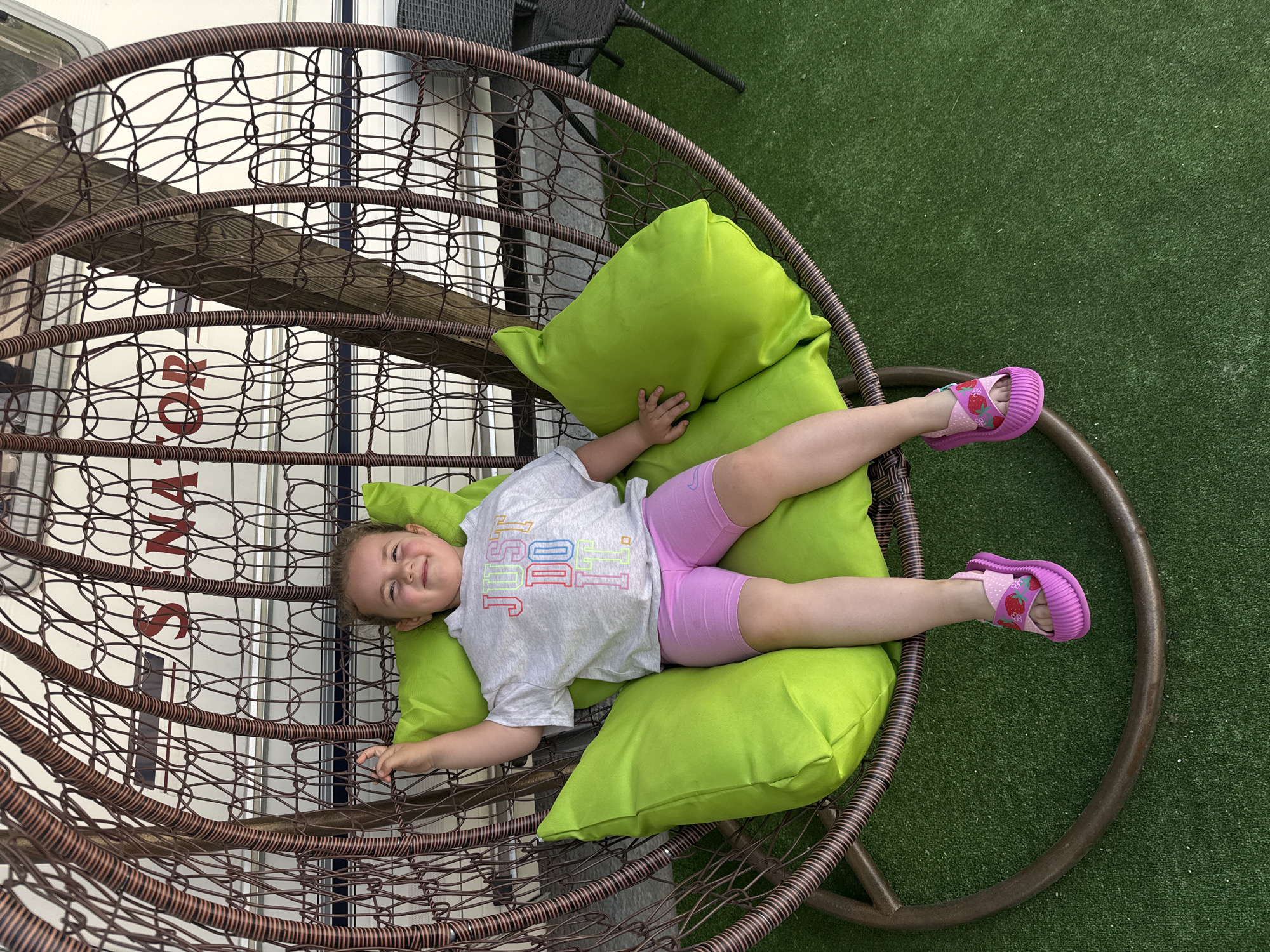 A young girl with blonde hair pulled back is seated in a hanging wicker chair with bright green cushions. She is wearing a light grey t-shirt with the words "JUST DO IT" in colorful lettering and pink biker shorts. Her feet are adorned with pink strawberry-themed sandals. The chair is suspended outdoors, with artificial green turf visible on the ground. In the background, a white caravan with red lettering reading "SENATOR" is partially visible. The scene appears to be set in a recreational area, possibly within a campsite or resort. The overall impression is one of a relaxed, sunny day.