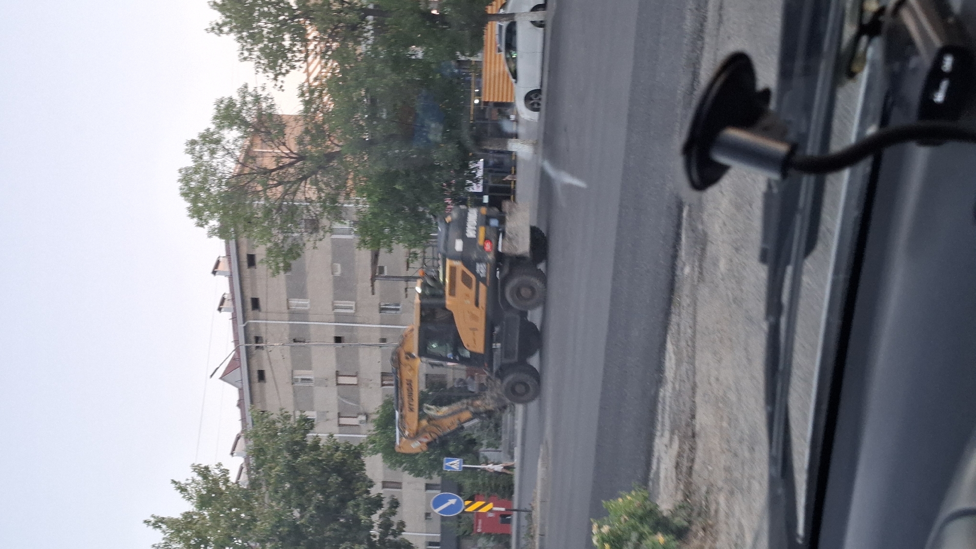 In Shymkent, Kazakhstan, an outdoor scene captures a yellow Hyundai wheeled excavator operating on a paved road. The excavator is positioned centrally in the frame, its large wheels visible on the asphalt. Behind the excavator, a multi-story apartment building with light-colored facade and numerous windows stands. Trees with green foliage flank the building and the road. A white sedan is parked on the right side of the road, beyond the excavator. Traffic signs are present on the left side of the road, including a blue circular sign with a white arrow pointing down and a white triangular sign with a black symbol. The sky above is a light blue, indicating daytime. The perspective suggests the photo was taken from inside a vehicle, with a dashboard and windshield wiper visible in the foreground.