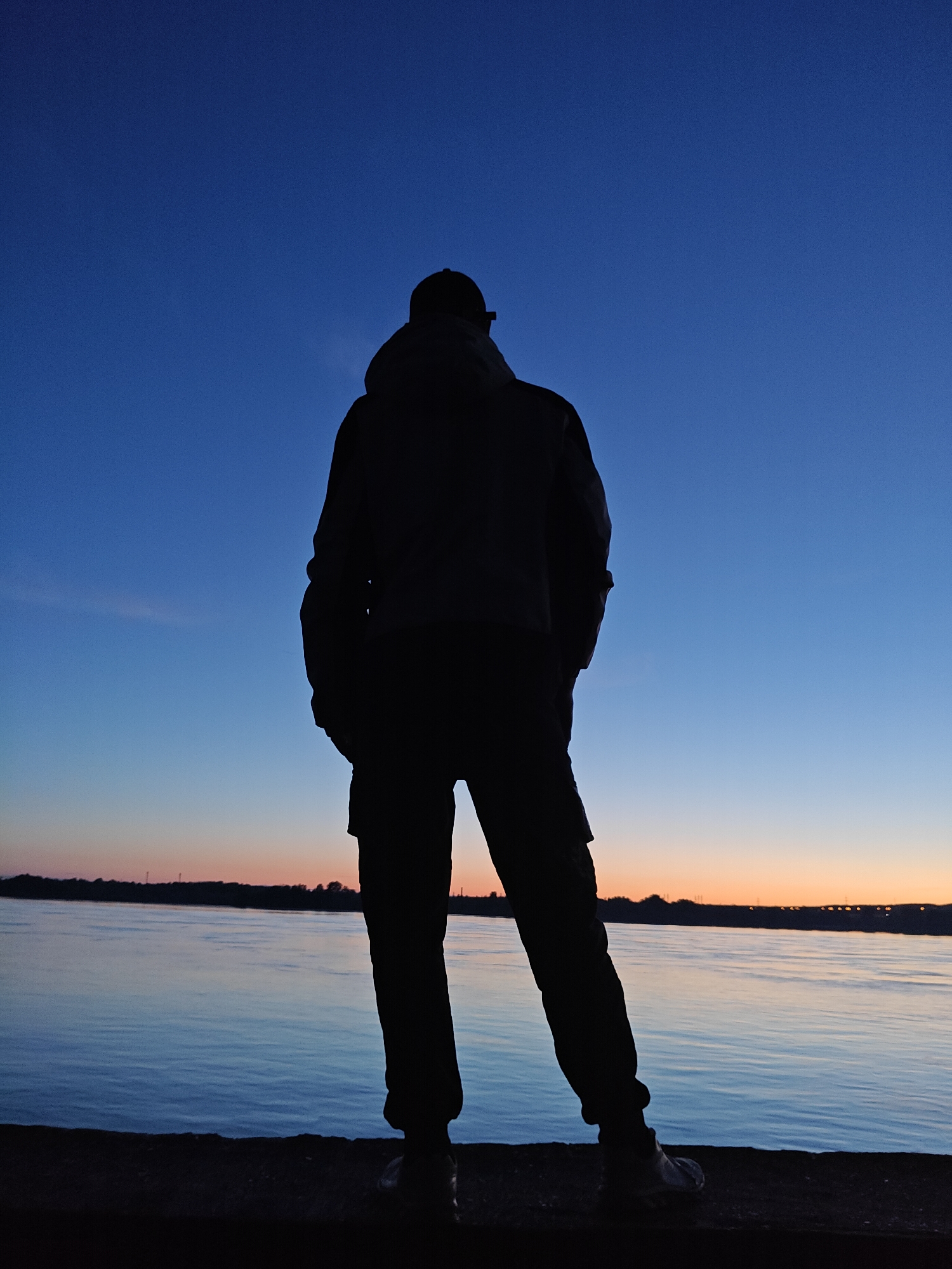 A lone individual stands in silhouette, viewed from behind, facing a wide body of water at what appears to be either twilight or dawn. The figure wears a cap, a jacket (possibly hooded), and pants, with hands in pockets, conveying a contemplative or observing stance.
The sky above displays a striking color gradient, transitioning from a deep, rich blue at the top, through a lighter azure in the middle, to warm orange and pink hues along the horizon. This suggests the last moments of sunset or the first light of sunrise. The large expanse of water, likely part of the Kama River or Votkinsk Reservoir in Chaikovsky, Russia, mirrors the sky's soft blues and oranges, exhibiting a calm, rippled surface.
In the distance, a dark, low-lying landmass marks the horizon, with faint, scattered lights or structures barely visible, hinting at a distant urban or industrial area. The person is standing on a dark, solid foreground surface, possibly a concrete embankment or pier. The overall scene evokes a sense of peaceful solitude and appreciation for the vast, changing sky and water. No text is visible in the image.