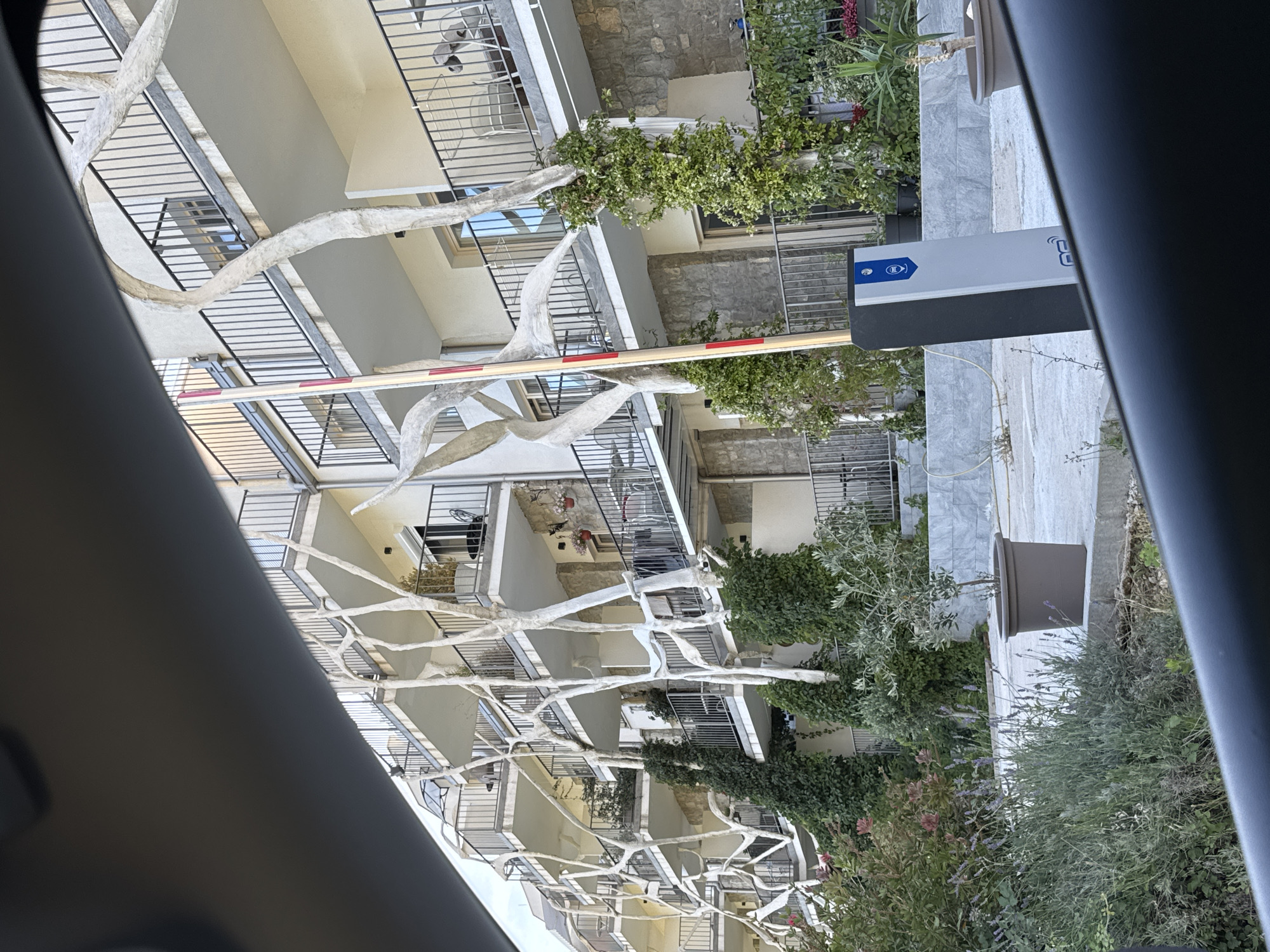 The image, taken from within a vehicle looking outwards, displays a multi-story building complex, likely a residential apartment building or hotel, under bright daylight. The building features several levels of balconies with black metal railings and a light-colored, possibly white or beige, facade.
A prominent feature in the foreground and midground is an intricate network of thick, white, branch-like structures that appear to be decorative elements, possibly trellises, attached to the building and spanning across multiple balconies. Some of these structures have green leafy plants growing on them.
At ground level, a red and white striped security barrier arm extends across what appears to be an access road or driveway, indicating controlled entry to the property. To the right of the barrier, a grey vertical post is visible, which seems to be part of the barrier system. This post has a blue and white logo or symbol near the top and partially visible text, possibly "COMM" or similar, on its side.
The immediate foreground includes part of the car's interior, dark in color, framing the shot. Beyond the barrier, there are various green plants and bushes, some with small flowers, adding to the lush outdoor environment. One large, brown potted plant is also visible on the tiled ground area. The architectural style, bright lighting, and vegetation are consistent with a Mediterranean climate, fitting the given location of Vrijesno, Montenegro. No people are visible, and no specific activity beyond the implied function of the security barrier is taking place.