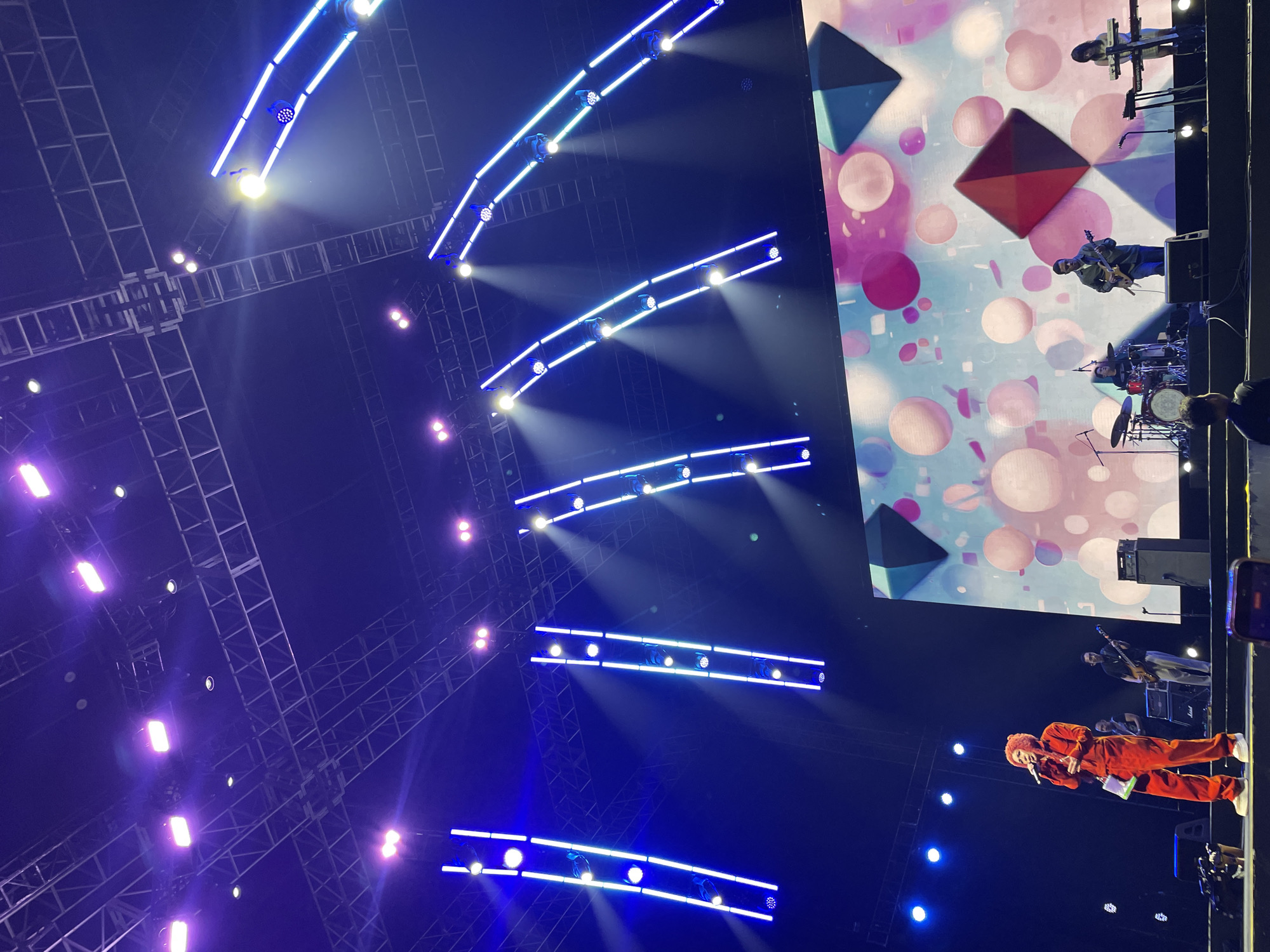 A low-angle shot depicts a live musical performance in Salembaran, Indonesia. In the foreground, a person in an orange jumpsuit holds a microphone. Stage lighting is prominent, with blue and white lights arranged in linear patterns overhead. A large screen displays a colorful, abstract design with geometric shapes and circles. Several band members, including guitarists and a drummer, are visible on the screen and partially on stage. The stage is set up with scaffolding and lighting rigs. Some concert attendees are standing in the lower corner of the frame. The lighting conditions are dim, emphasizing the stage lights.