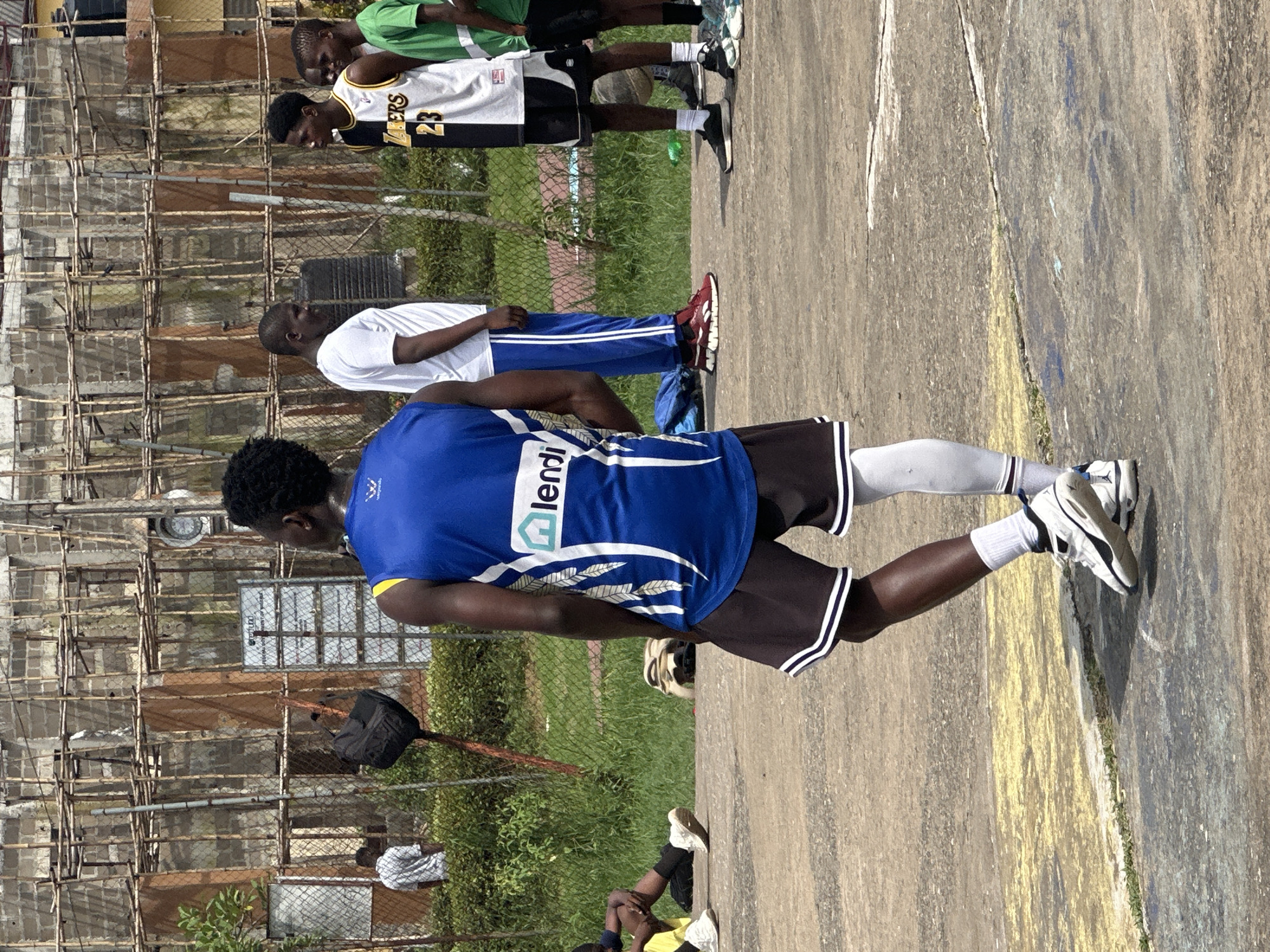 In Ajara, Nigeria, on a sun-drenched outdoor basketball court with a weathered concrete surface marked with faded yellow lines, a group of young men are engaged in activity. The court is bordered by a chain-link fence, beyond which lies overgrown green grass and a multi-story building under construction, its exterior a patchwork of concrete blocks and scaffolding.
In the foreground, a young man wearing a blue basketball jersey with white trim and a "lendl" logo, and dark shorts with white stripes, is captured from the rear, his body angled away from the camera. He sports white knee-high socks and white basketball shoes with blue accents. To his right, another young man is dressed in a black and white basketball jersey labeled "LAKERS 23" and black shorts, also wearing white socks and black basketball shoes. Further to the right, a third individual is visible in a green t-shirt and black shorts.
In the middle ground, a person in a white t-shirt and blue pants with white stripes stands facing the group. Another figure in a white shirt stands with their back to the viewer on the far left, near the fence. A black backpack is hanging from a pole near the construction site. The overall atmosphere suggests a casual basketball game or practice session in a semi-urban Nigerian setting.