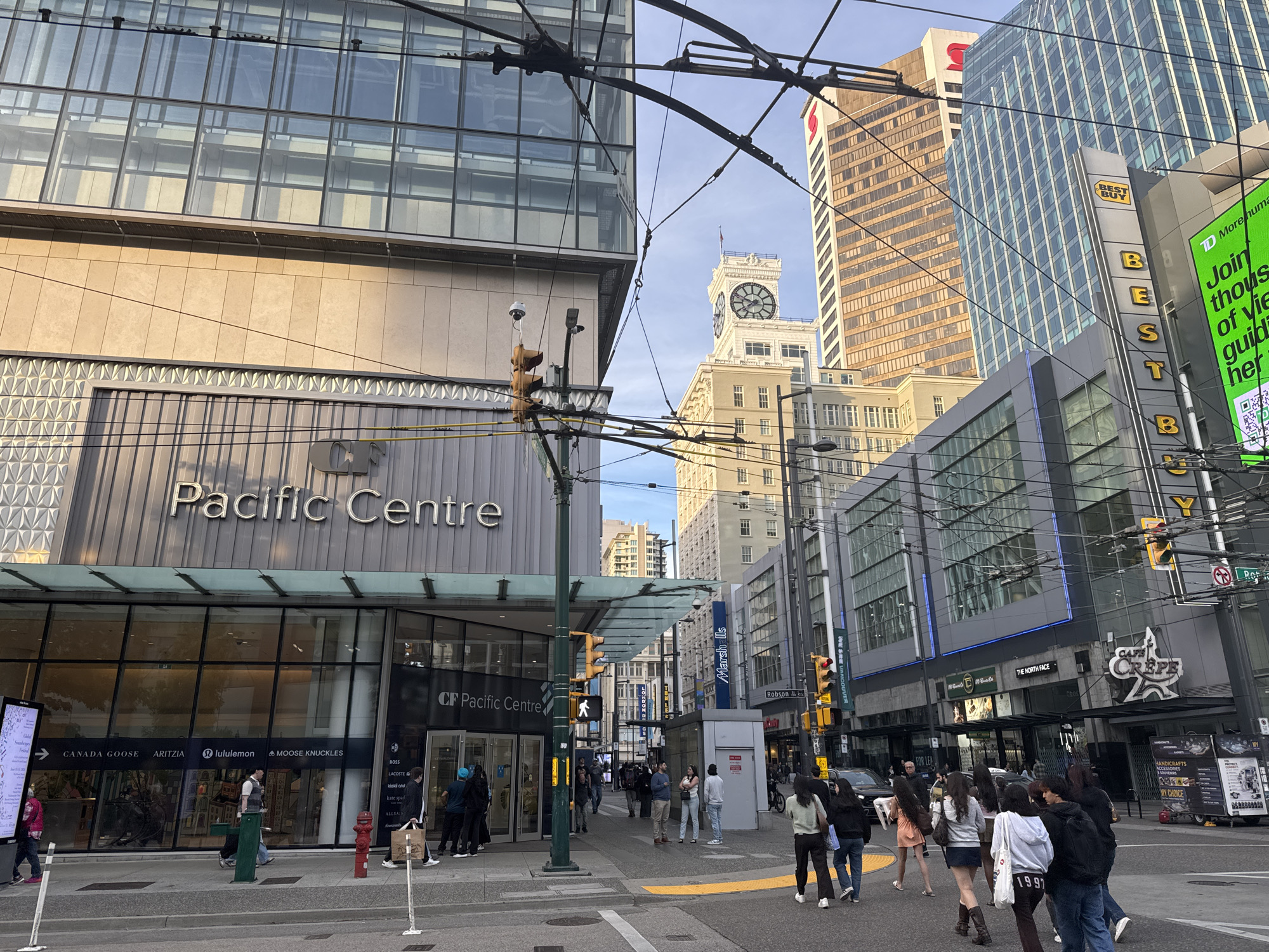 This image depicts a street scene in downtown Vancouver, Canada. It showcases the "Pacific Centre" shopping mall prominently on the left side of the frame. The building features a modern architectural design. A variety of retail stores, including Canada Goose, Aritzia, Lululemon, and Moose Knuckles are visible behind the storefront windows. A pedestrian crosswalk is shown in the foreground with a group of people crossing the street. A pedestrian traffic light stands at the crosswalk. The "Best Buy" logo is visible in the background on a building to the right. A building with a clock tower is also visible in the distance. The sky is mostly clear with a few clouds. Overhead trolley lines crisscross the street.