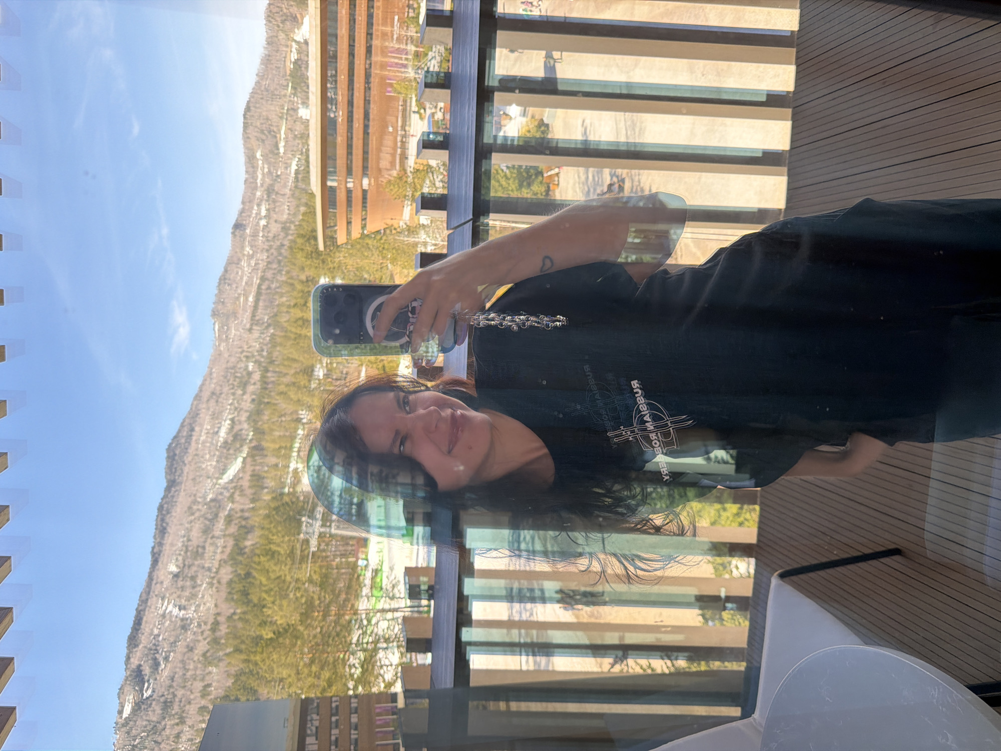 A woman is taking a selfie with a smartphone against the background of a mountain and a building in a city, possibly Озёрное, Russia. The view is seen through a glass window with multiple reflections. The woman is wearing a dark-colored shirt and a necklace. The city in the background contains buildings, trees, and a clear sky.
