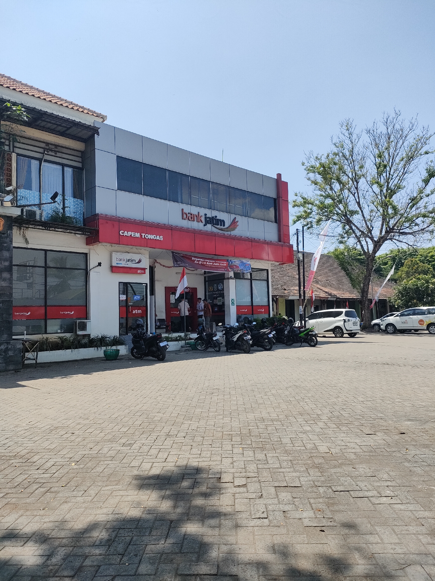 The image shows the exterior of a Bank Jatim branch located in Curahdringu, Indonesia. The building features a modern facade with prominent red and gray signage for "bank jatim" and "CAPEM TONGAS". To the left of the main building, there is an older structure with a tiled roof and windows. In front of the bank, a paved area is occupied by several parked motorcycles. To the right of the building, two white cars are parked, one with "Jatim" branding, and beyond them, traditional Indonesian houses with tiled roofs and trees are visible. The sky is clear and blue, suggesting a sunny day. A few people are visible near the entrance of the bank.