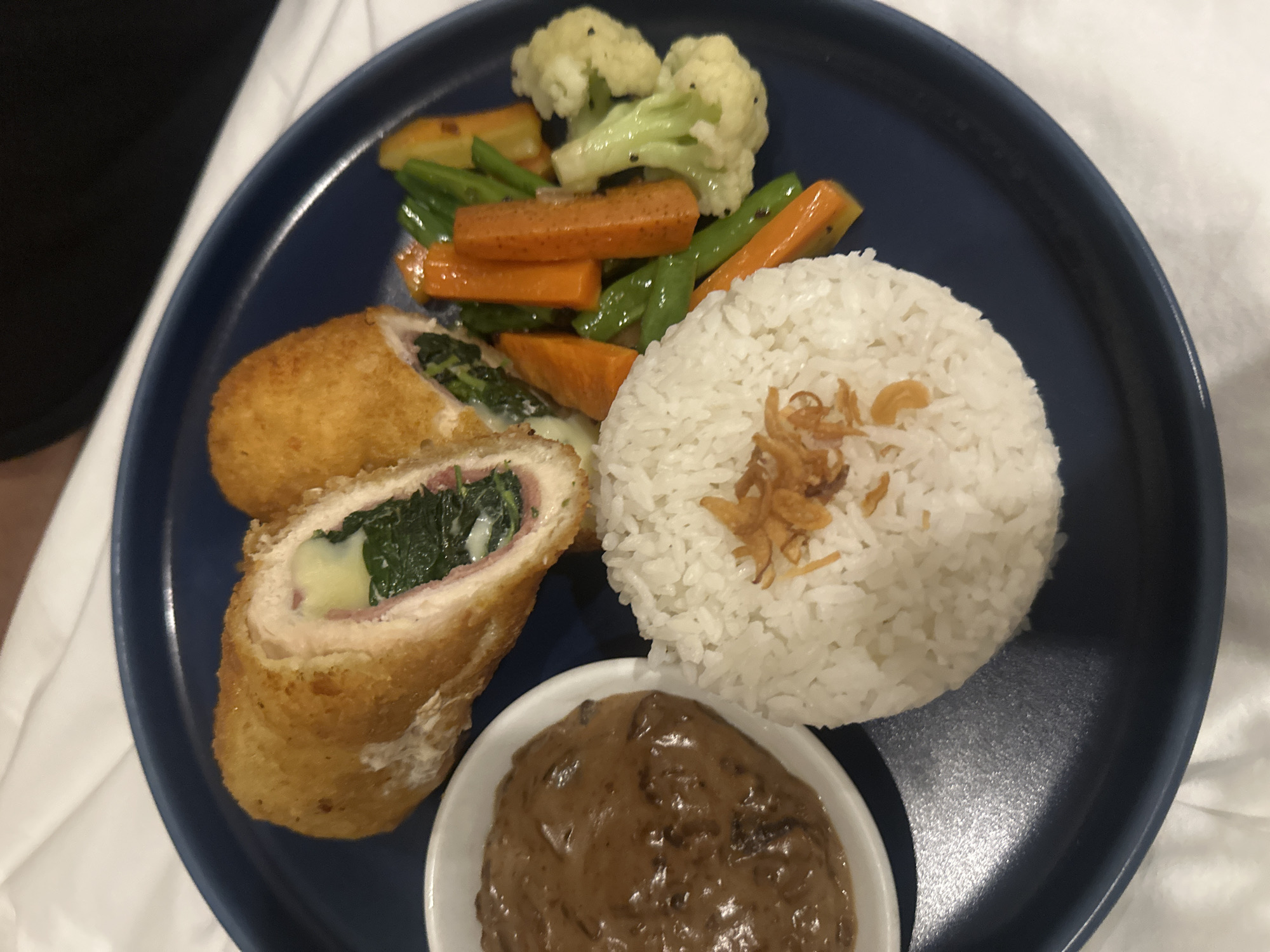 A well-presented meal is captured on a dark blue, round plate, set against a soft white background, likely a tablecloth or bed linen. The plate features a substantial main course comprising several distinct elements.
The focal point is a breaded and fried chicken roulade, possibly a chicken cordon bleu or similar dish. One piece is sliced open, revealing a vibrant green spinach filling, melted white cheese, and pinkish processed meat (likely ham), all encased in a golden-brown, crispy breading.
To the right of the chicken, a neat mound of fluffy white rice is garnished with crispy fried shallots or onions, adding a textural and aromatic contrast. Above the rice and chicken, a colorful medley of steamed or sautéed vegetables is arranged. This includes florets of white cauliflower and green broccoli, bright orange carrot sticks, and slender green beans, some with small dark specks, possibly seasoning.
A small, white, circular ramekin holds a creamy, rich brown sauce, resembling mushroom gravy, positioned at the bottom right of the plate. The overall presentation is tidy and appetizing, suggesting a prepared meal served perhaps in a restaurant or hotel setting in Kuta Selatan, Indonesia. No visible text is present in the image.