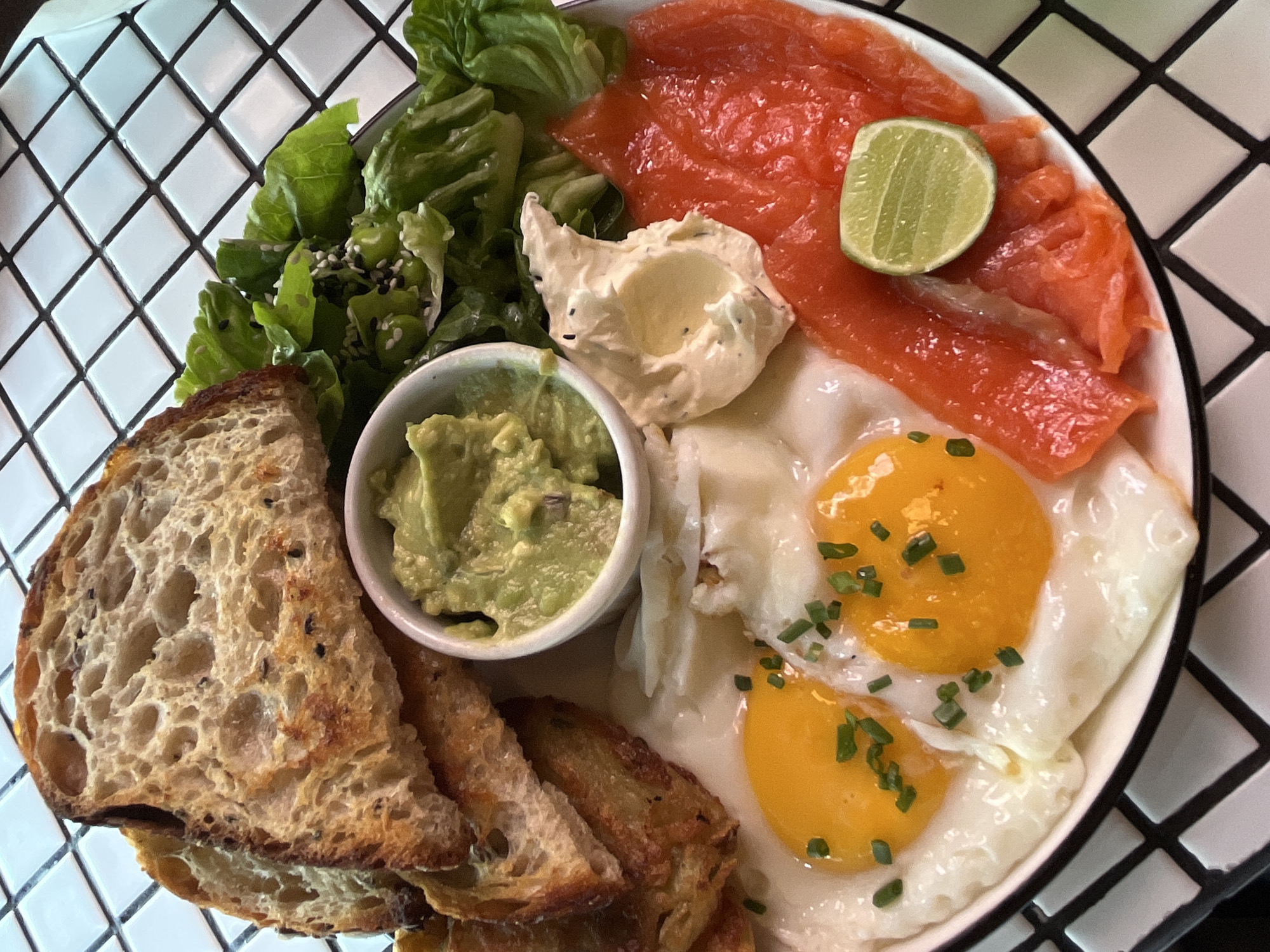 A close-up, eye-level shot captures a visually appealing breakfast plate set against a black and white tiled surface. The plate contains a variety of food items: two fried eggs with bright yellow yolks, garnished with chopped chives; thinly sliced smoked salmon; a small dollop of a creamy, light-colored spread, possibly cream cheese or aioli, with visible herbs; a small white ramekin filled with mashed avocado; a pile of toasted bread slices with a textured crust; and a portion of green salad with visible sesame seeds and edamame. A single lime slice rests on the salmon. The overall presentation suggests a well-prepared and healthy meal. The location is specified as North Kuta, Indonesia.