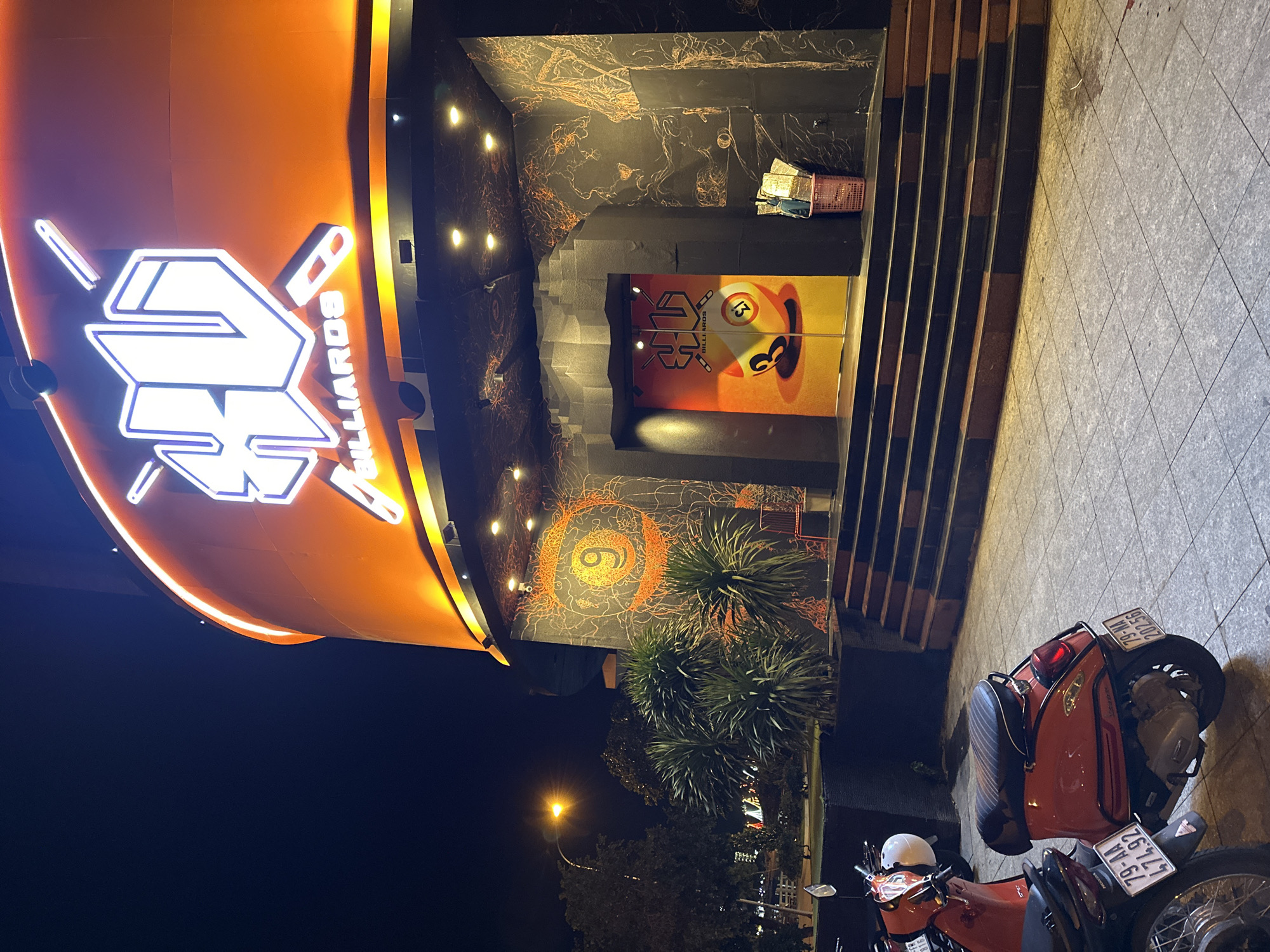 The image depicts the exterior of a billiards establishment in Nha Trang, Vietnam, at night. The building features a prominent, large, orange, circular facade illuminated by neon lights, displaying the text "XV BILLIARDS." Below the main sign, a dark, textured entrance is visible, flanked by a staircase leading up to it. To the left of the entrance, a large, orange circular mural with the number "9" in the center is displayed on the wall. To the right of the entrance, a pink laundry basket is filled with items, including what appear to be bags and possibly clothing. Two motorbikes are parked in the foreground on the tiled pavement. The one on the left is partially visible, showing a license plate "79-MA 474.92". The motorbike on the right is a red scooter, also with a visible license plate "79-MA 202.56". The overall lighting suggests it is nighttime, with streetlights and building lights illuminating the scene.