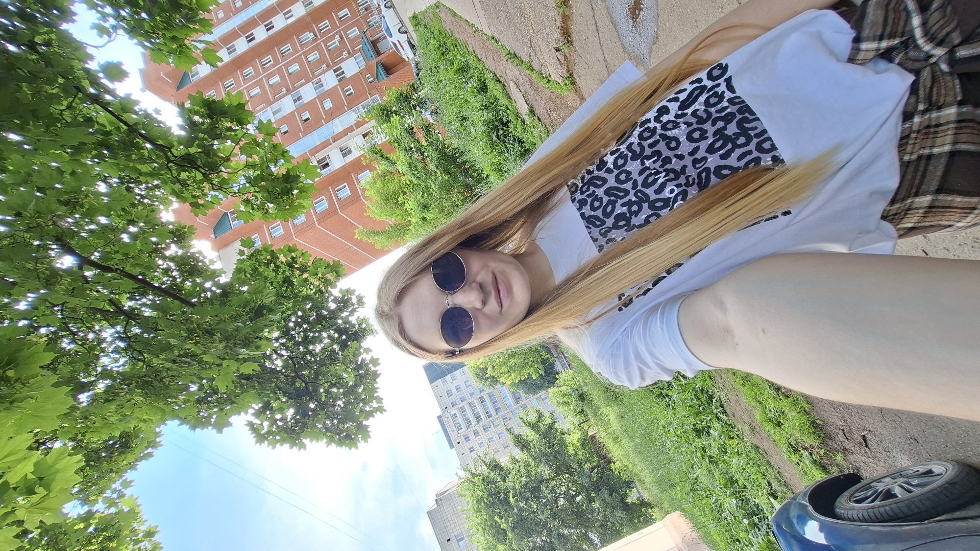 In a wide-angle selfie taken outdoors, a young woman with long blonde hair and round sunglasses stands on a paved pathway. She is wearing a white t-shirt with a leopard print design on the front and a plaid skirt tied around her waist. Behind her, a green, leafy tree partially obscures the view of a tall, multi-story brick apartment building with many windows. To her left, another apartment building, this one lighter in color and with a more uniform facade, is visible, surrounded by green grass and trees. A dark-colored car is parked on the left side of the frame. The sky is bright blue with scattered white clouds, and the sun is casting light through the tree's leaves. The scene appears to be a residential area in the city of Chaikovsky, Russia, possibly during the daytime in warmer weather, given the lush greenery and light clothing.
