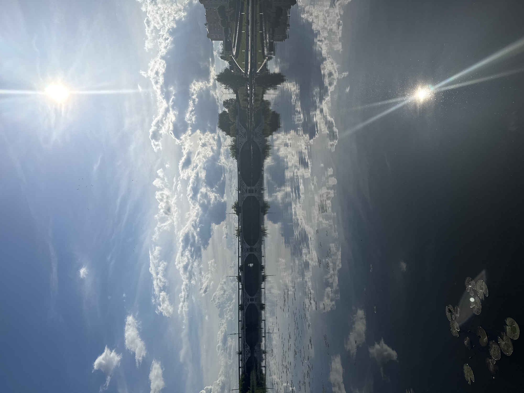 The image shows a reflection of a bridge in a body of water, possibly a pond or lake. The bridge structure is visible in the center, with a symmetrical reflection beneath it. The sky above is blue with scattered white clouds, and the sun's reflection is visible on either side. Lily pads are scattered on the water's surface. Given the context provided, the image likely depicts the Votkinsk Pond bridge located in Votkinsk, Russia.