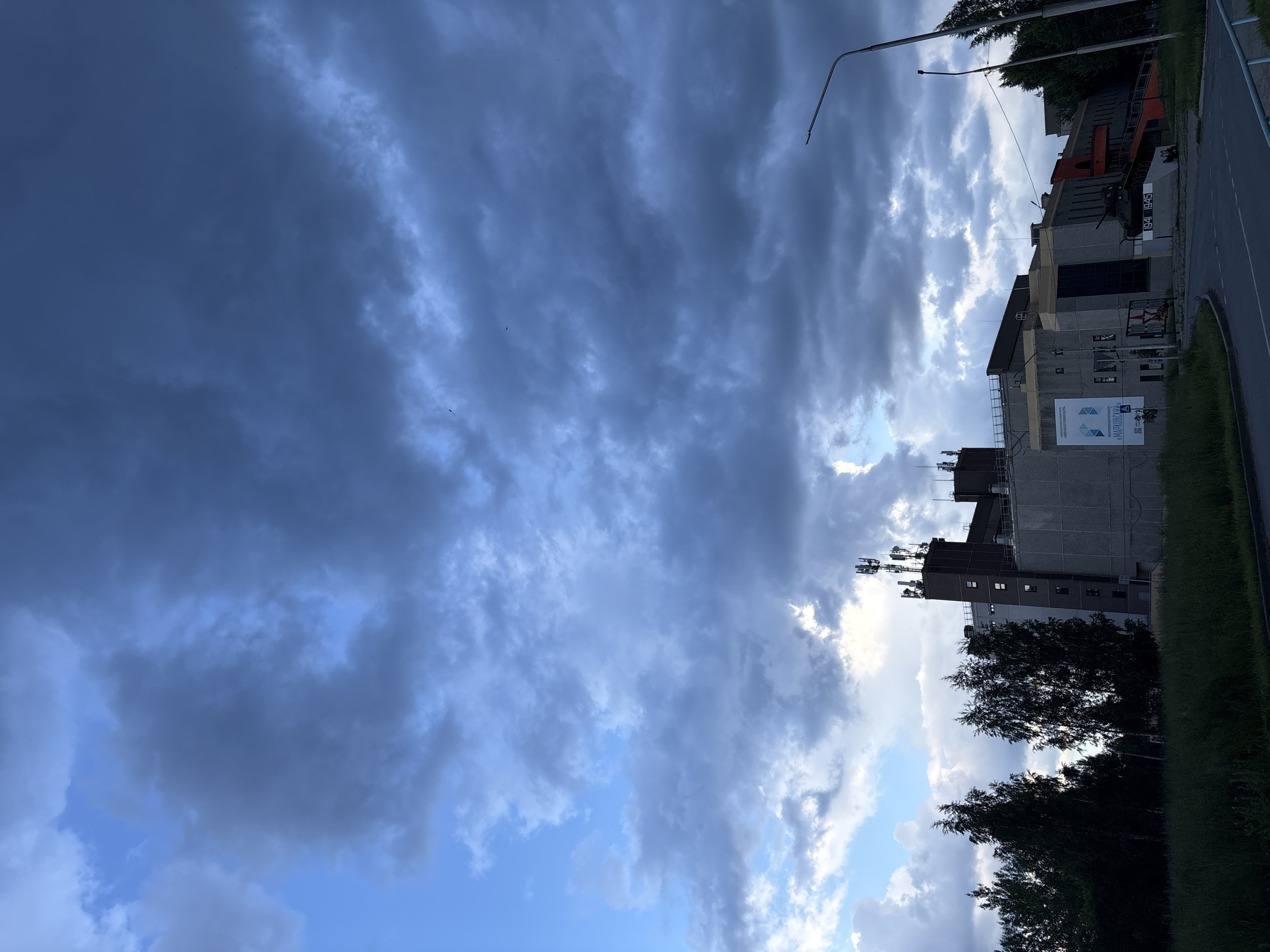 The image captures an outdoor scene dominated by a dramatic, cloudy sky with pockets of blue visible. In the mid-ground, a large, multi-story building complex with concrete facades and several antennae on its roof is visible. A portion of the building's facade appears to be under construction or covered with sheeting. To the left of the building, a cluster of dark green trees stands out against the lighter sky. A street runs in the foreground, with a lamppost on the right side of the image. To the left of the building, a small display with text and an image is mounted on the wall, and a red star insignia is visible on another section of the building. In front of the building, a military tank is displayed on a pedestal with the dates "1941 1945" inscribed on it. The scene is likely in the city of Марковский, Russia, given the context.