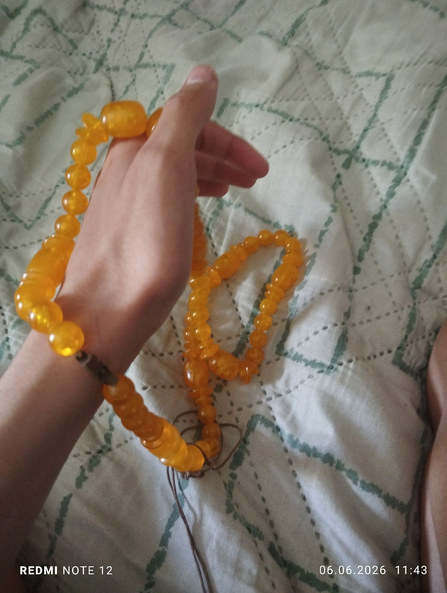 The image depicts an indoor scene in Burgas, Bulgaria. A single human left hand with a light skin tone is prominently featured, holding a strand of amber-colored prayer beads (tasbih or misbaha). The beads are translucent, yellow-orange, and appear to be irregular in shape, strung on a visible dark brown cord. A distinct, larger "imam" bead is visible near the thumb. An additional, unheld segment of similar beads is coiled on the surface to the right of the hand. The beads and hand rest upon a light-colored, likely white or cream, quilted fabric surface, identifiable as a blanket or bedspread. This fabric exhibits a distinct pattern of intersecting lines and dashed stitches, highlighted with a faded teal or light green thread. A portion of a human limb, consistent with a leg or foot, is visible in the lower right corner of the frame. Overlays in the bottom left corner identify the device as "REDMI NOTE 12," and in the bottom right, the date "06.06.2026" and time "11:43" are displayed.
