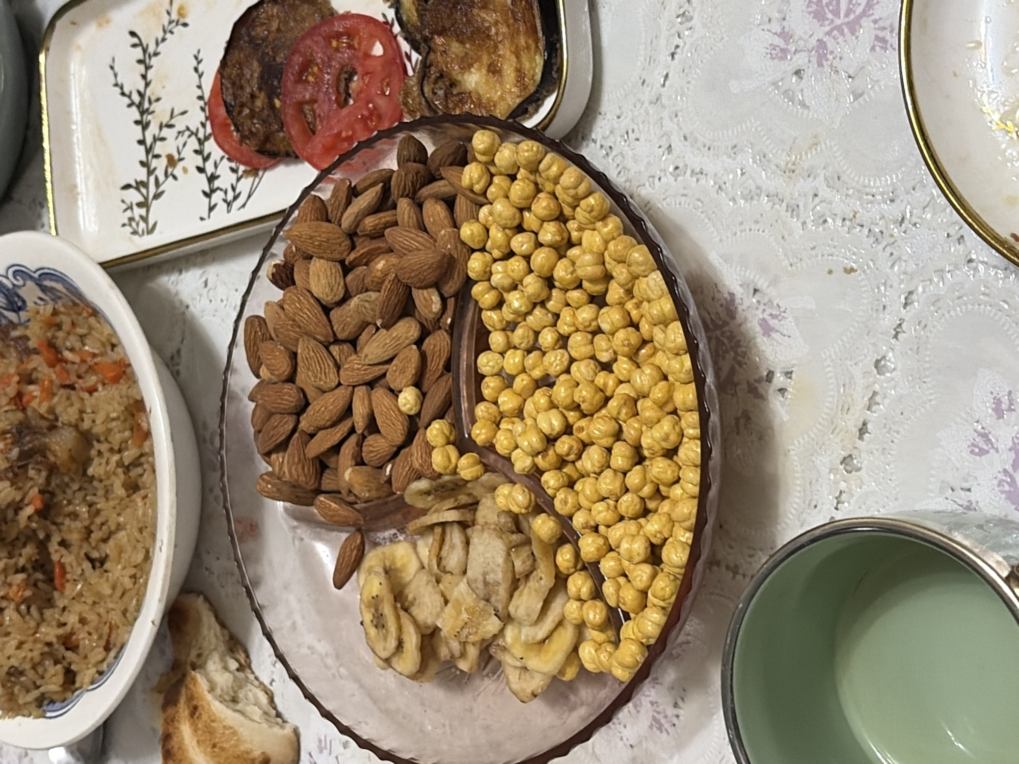 The image shows a close-up, overhead view of a table laden with food, likely a meal or gathering. The setting appears to be indoors, with a white, patterned tablecloth.
Dominating the center of the frame is a clear, shallow, circular dish containing a variety of snacks. On the left side of the dish are numerous whole almonds. To the right of the almonds are bright yellow, round, roasted chickpeas. At the bottom of the dish, in a smaller section, are dried banana chips.
To the left of the snack dish is a white bowl filled with a savory rice dish, appearing to be pilaf, with visible pieces of meat and carrots. Next to the rice bowl, a piece of crusty white bread is visible.
In the upper left corner, a white rectangular plate holds food items including what looks like a fried cutlet or patty, topped with slices of red tomato and accompanied by pieces of what might be fried eggplant. Another white plate with a gold rim is partially visible on the right side of the image, suggesting it's part of the same meal setting. A light green ceramic mug or pot is also present in the lower right corner.
The lighting suggests daytime, possibly late afternoon or evening, given the natural light that seems to be illuminating the scene. There are no people visible in the image, and the focus is entirely on the food. The overall impression is one of abundance and a shared meal. There is no visible text in the image.