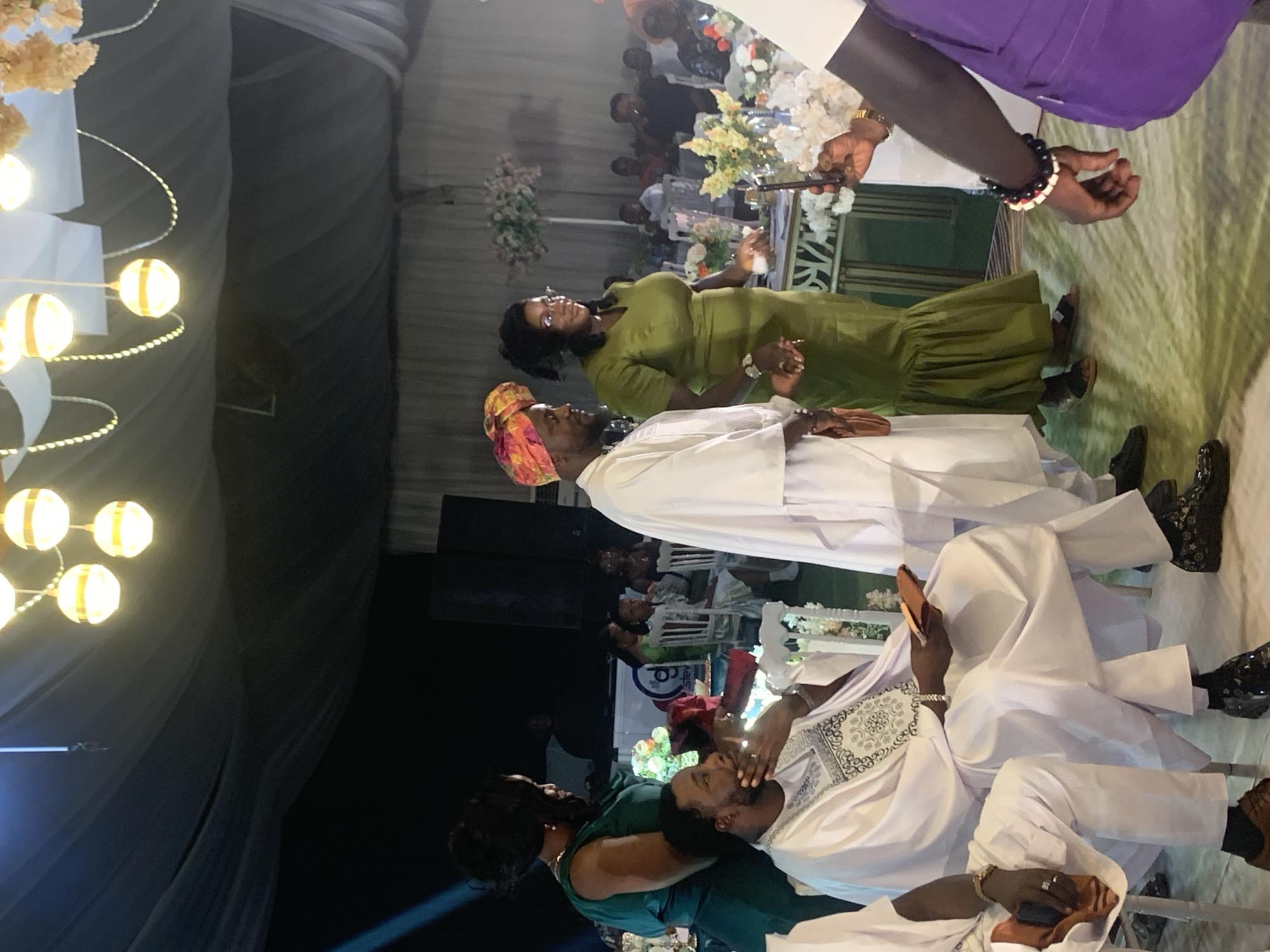 This image depicts an indoor social gathering, likely an evening event, characterized by decorative lighting and floral arrangements. The setting appears to be a large tent or event hall, indicated by the draped fabric ceiling and walls.
In the immediate foreground, a man wearing a white kaftan with intricate embroidery is seated, resting his head on the shoulder or lap of a woman in a dark green dress, who is seen from behind. The man in the kaftan wears brown shoes and holds a phone in one hand. Standing near them is another man dressed in a white kaftan and a vibrant, multi-colored headwrap, wearing black shoes. He is facing the seated couple, appearing attentive. To his right, a woman in a long olive green dress and glasses stands, holding a glass and also looking towards the seated pair.
In the upper right portion of the frame, a person wearing a purple shirt or jacket is partially visible, holding a long, thin object that might be a microphone or a decorative item, above a table adorned with white and cream floral arrangements. Other guests are seated at tables further in the background, illuminated by soft lighting. The venue is decorated with glowing orb-shaped string lights suspended from the ceiling. The attire worn by the subjects, particularly the kaftans and headwrap, suggests a cultural event in Nigeria, consistent with the location context of Jos. The scene conveys a relaxed yet engaged atmosphere among the attendees. No discernible text is visible in the image.