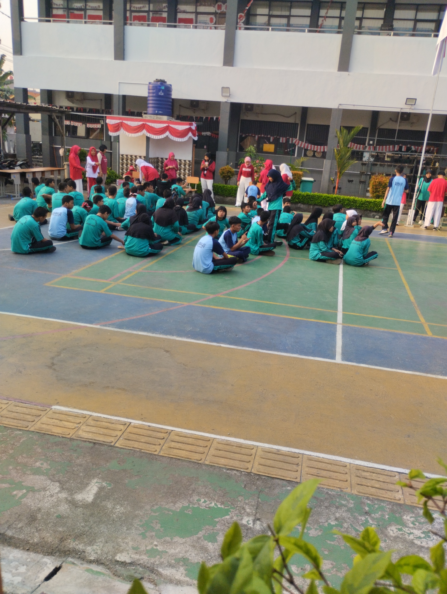 In Bekasi, Indonesia, a group of students in teal and white uniforms are gathered on a sports court. Many are sitting cross-legged, facing a raised platform adorned with red and white bunting and draped fabric. Several adults, some in similar uniforms and others in red attire, stand near the platform, appearing to address or direct the students. The sports court has painted lines in blue, yellow, and green, and a portion of a building with multiple windows is visible in the background. Some students are interacting with each other, while others are listening intently. The scene suggests an outdoor school event or assembly.