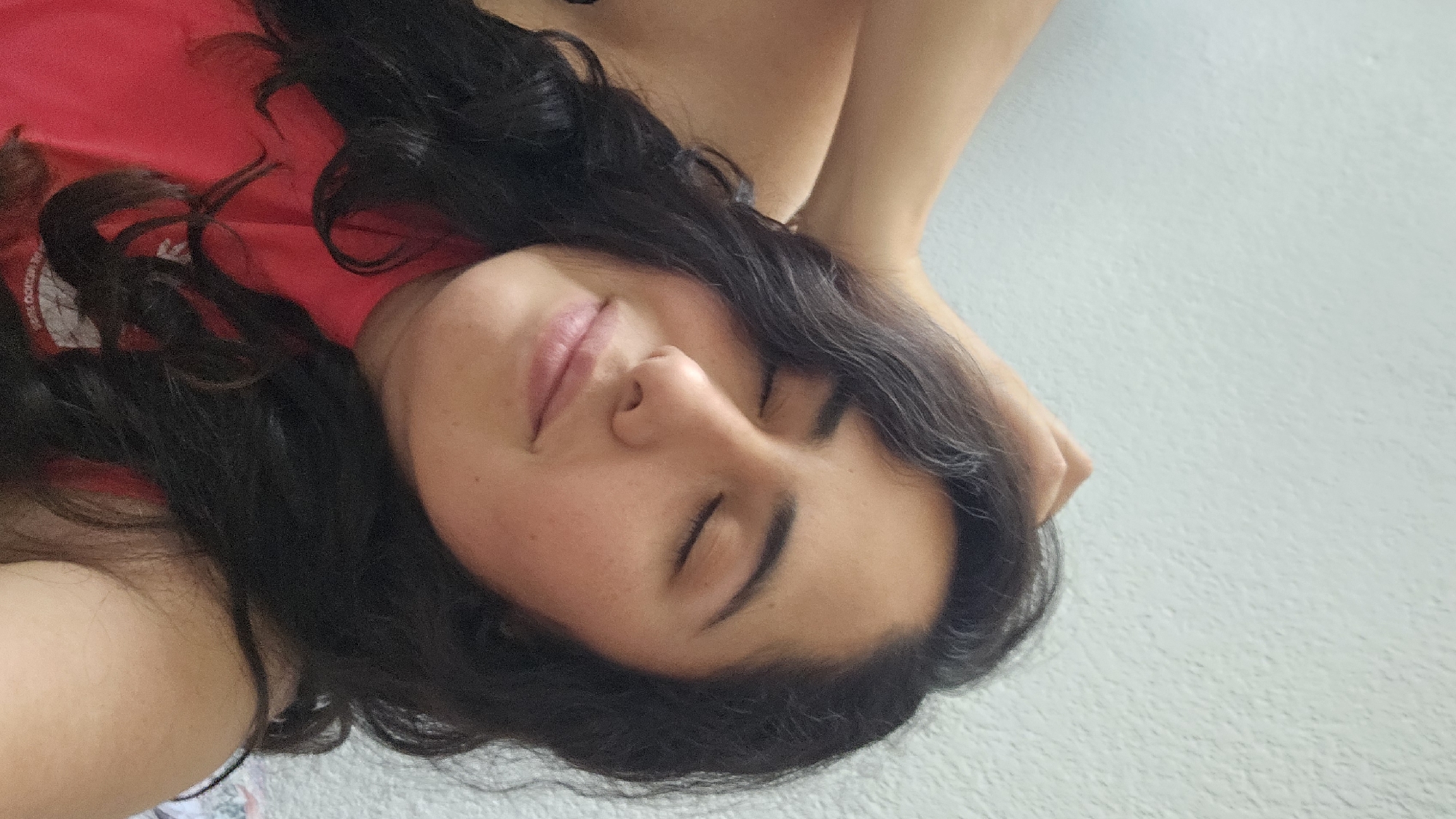 A woman with dark, wavy hair and closed eyes is lying down. She is wearing a red shirt. Her skin appears fair with some freckles. Her right arm is raised near her head, which rests on a surface or object. The background is a plain, light-colored wall with some texture. Without further information, the location in San Bernardino Tlaxcalancingo, Mexico, cannot be confirmed.