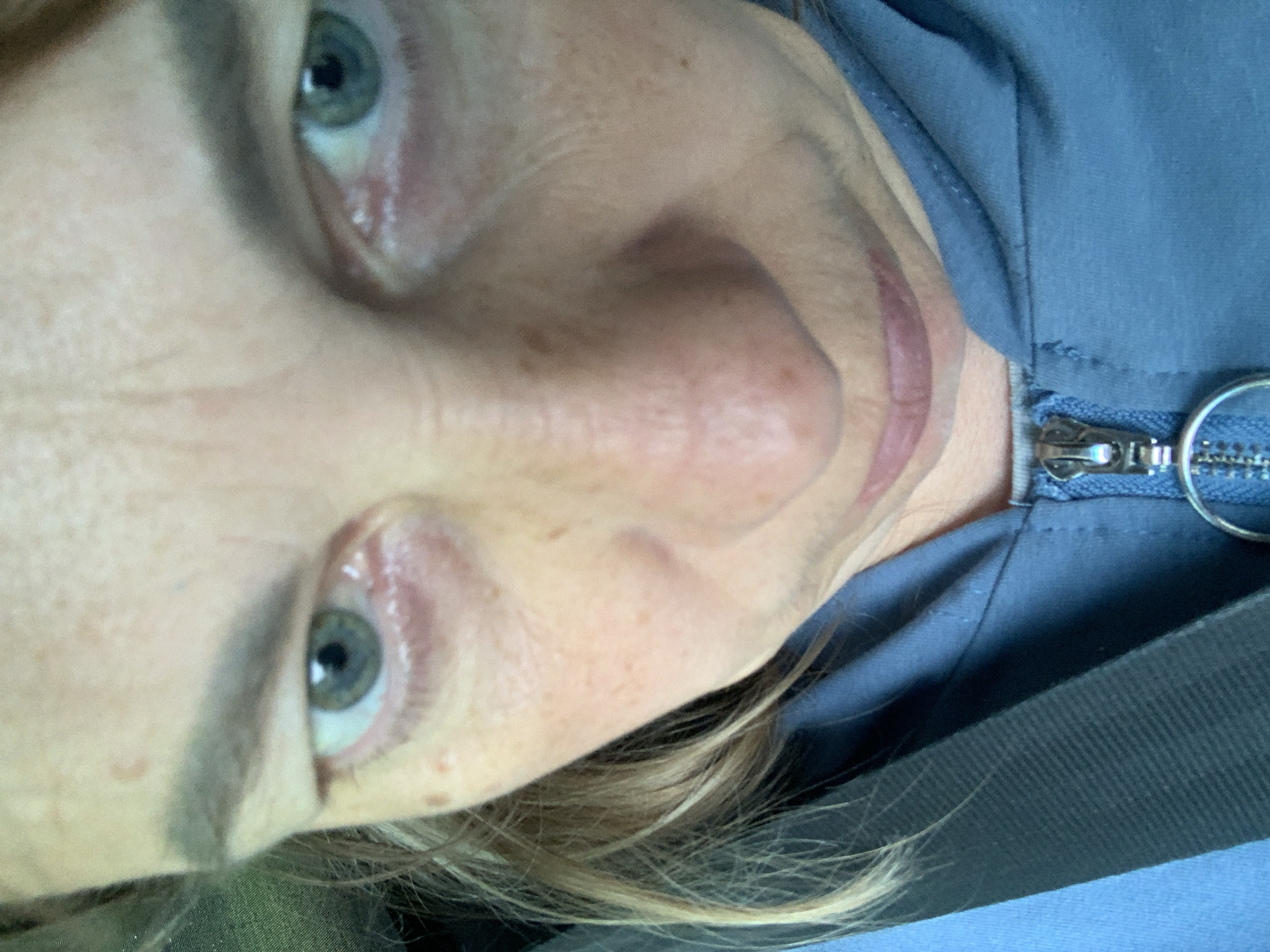 A close-up, high-angle shot shows a person's face, predominantly the eyes, nose, and mouth, from the nose up. The individual has fair skin with freckles and light brown hair. They are wearing a blue zippered jacket. The lighting suggests an outdoor or well-lit indoor environment. The provided location, Мядзельскі сельскі Савет, Belarus, is a rural administrative unit, implying the photo was taken in a village or rural area within that region. The scene captures a moment of personal photography, possibly a selfie, with no other individuals or specific environmental details of the location visible.