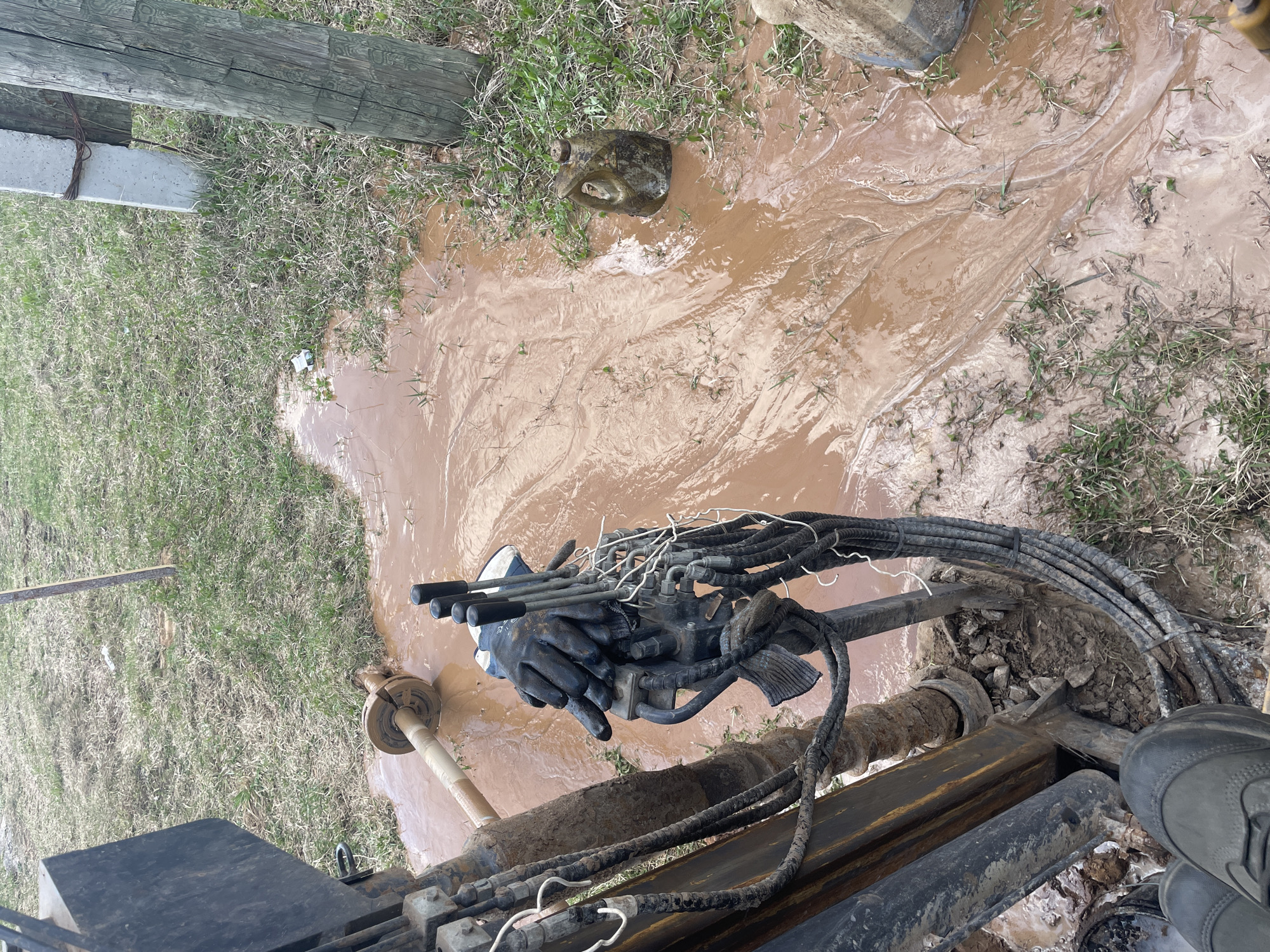 A top-down perspective shows a muddy area surrounding a drilling rig. Brown, sediment-laden water covers much of the ground, flowing around patches of grass. The drilling rig's hydraulic controls and hoses are visible, covered in mud, with one hand wearing a black glove operating the levers. A small, dark-colored plastic container sits on the edge of the muddy area, partially obscured by grass. To the left, a portion of a wooden fence post is visible. The environment appears to be a rural or undeveloped area. The city of Поедуги, Russia is not evident in the image.