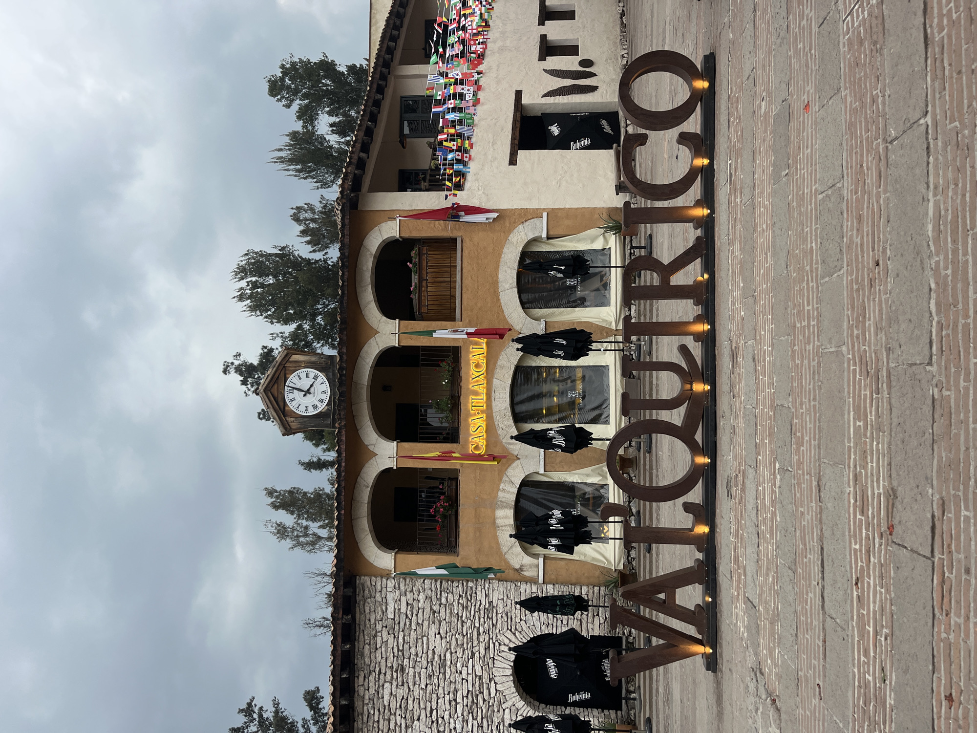 The image shows the facade of "Casa Tlaxcala" in Domingo Arenas, Mexico. The building has a clock tower, three levels with arched windows, and is decorated with national flags of different countries. A large metal sign spelling "VALQUIRICO" stands in front of the building. The sky is overcast. The ground is paved with brick. Several black umbrellas are open near the building's entrance.