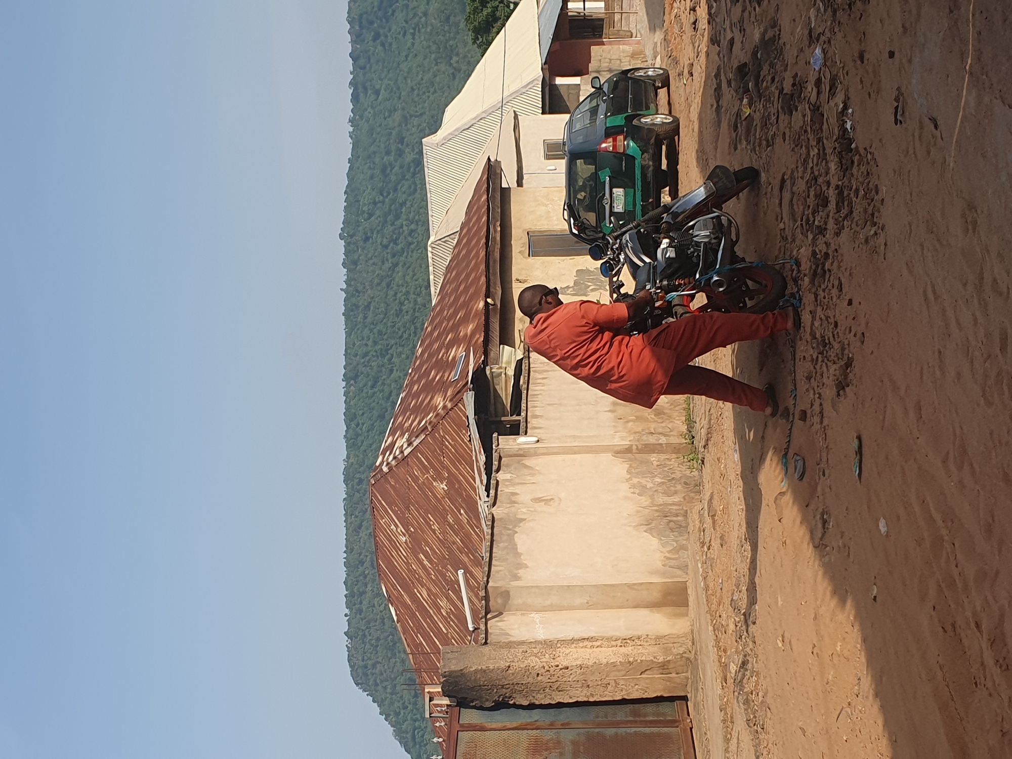A man in an orange outfit is standing in a dusty, outdoor area next to a motorcycle. The motorcycle is dark colored with silver accents. The man appears to be adjusting or working on the motorcycle, possibly securing it with a rope or chain. In the background, there is a weathered building with a rusty, corrugated metal roof. Further in the background, there is a dark green SUV parked on a dirt road. Beyond the buildings, a lush, green, hilly landscape is visible under a clear blue sky. The scene appears to be in a residential or semi-urban area within Lokoja, Nigeria.