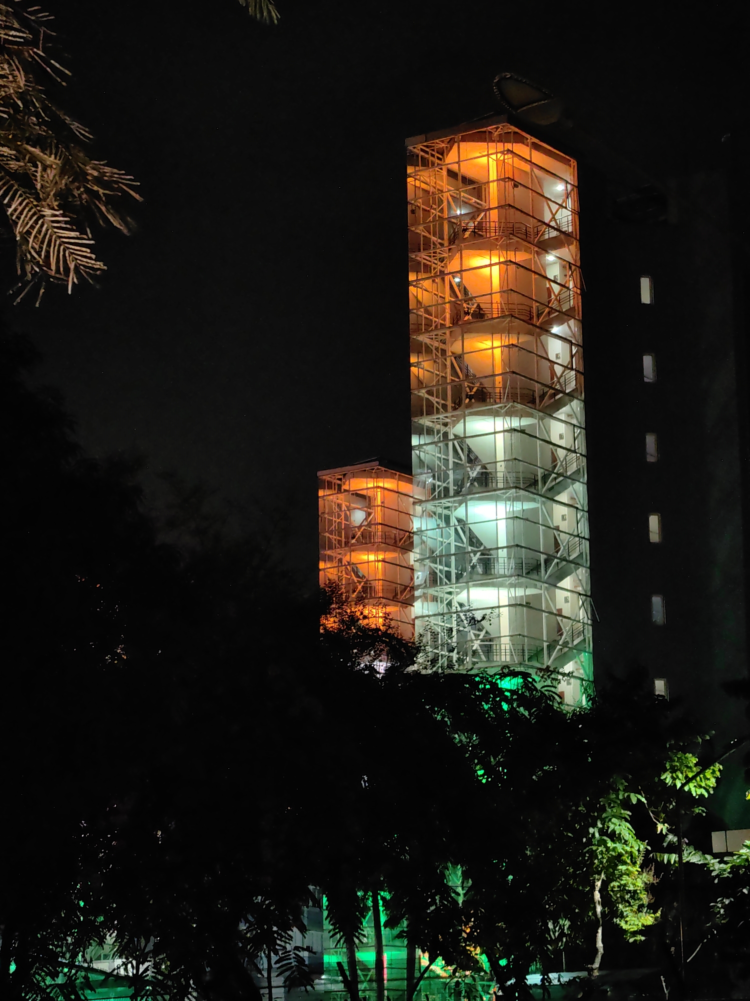 The image shows a night view of a multi-story building with illuminated sections in shades of orange and green. The building is a tall, slender structure with a visible framework and glass panels. The orange lights are concentrated in the upper portions of the structure, while the lower sections are lit with green. To the right of this building, another darker building with rectangular windows is visible. In the foreground, silhouetted trees and foliage obscure the lower parts of the illuminated buildings, with some green lights visible through the leaves. The overall scene suggests an urban environment at night, with the building's lighting possibly indicating a special occasion or a themed display. The description states this is located in Mumbai, India.